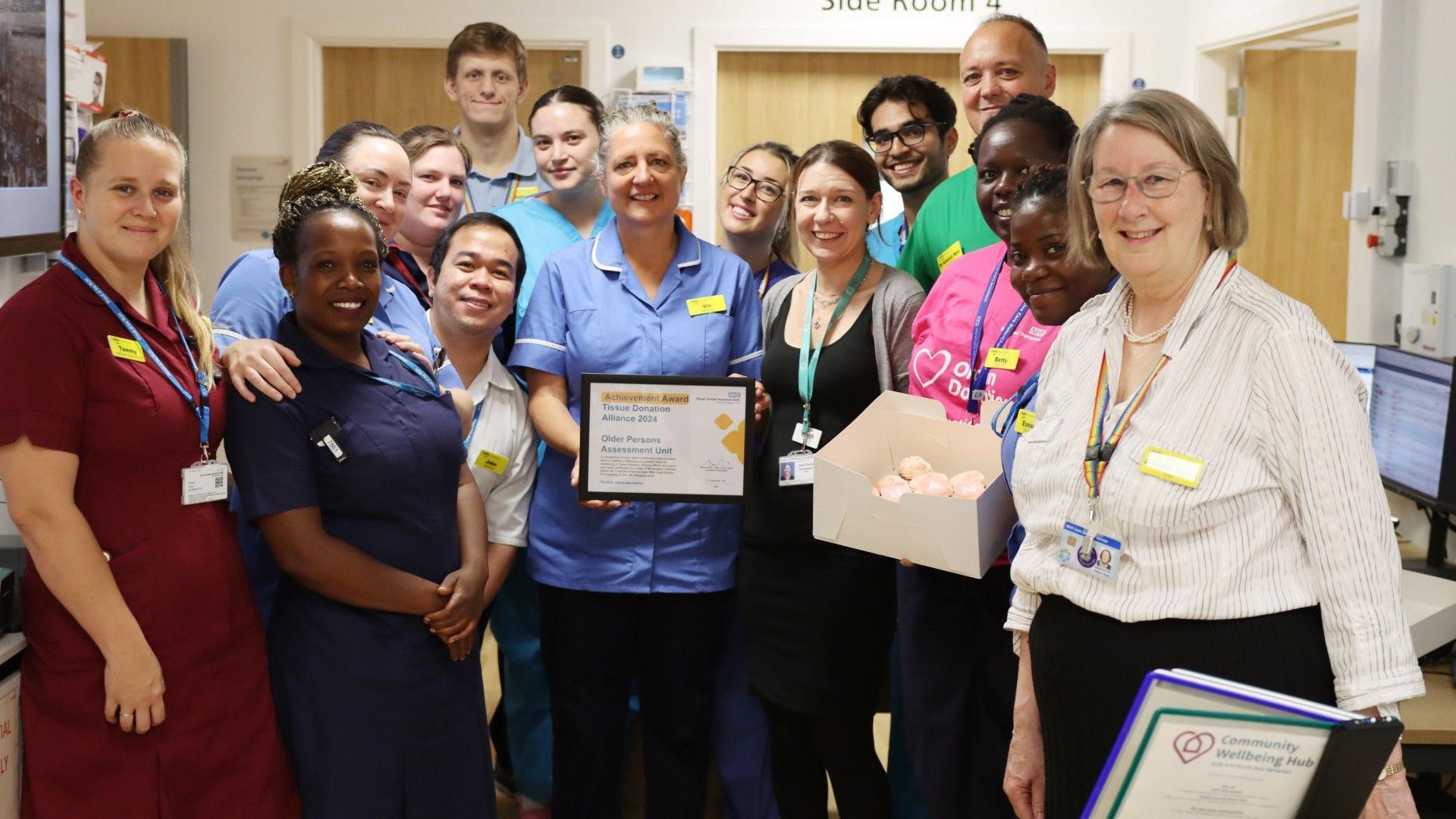 A group of medical staff at RUH Bath. They are all pictured standing close together in a group, smiling at the camera. Many of them are wearing scrubs, but one woman is wearing a pink Organ Donation t-shirt. One woman is holding a framed certificate and another is holding a box of pink cakes. 