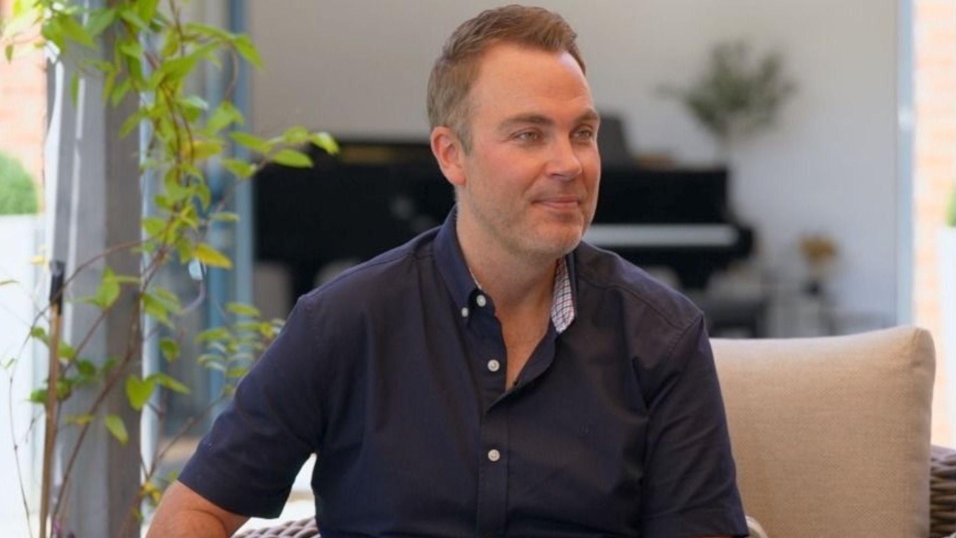 A man sitting on a chair inside, wearing a blue short sleeve shirt. There is a piano behind him
