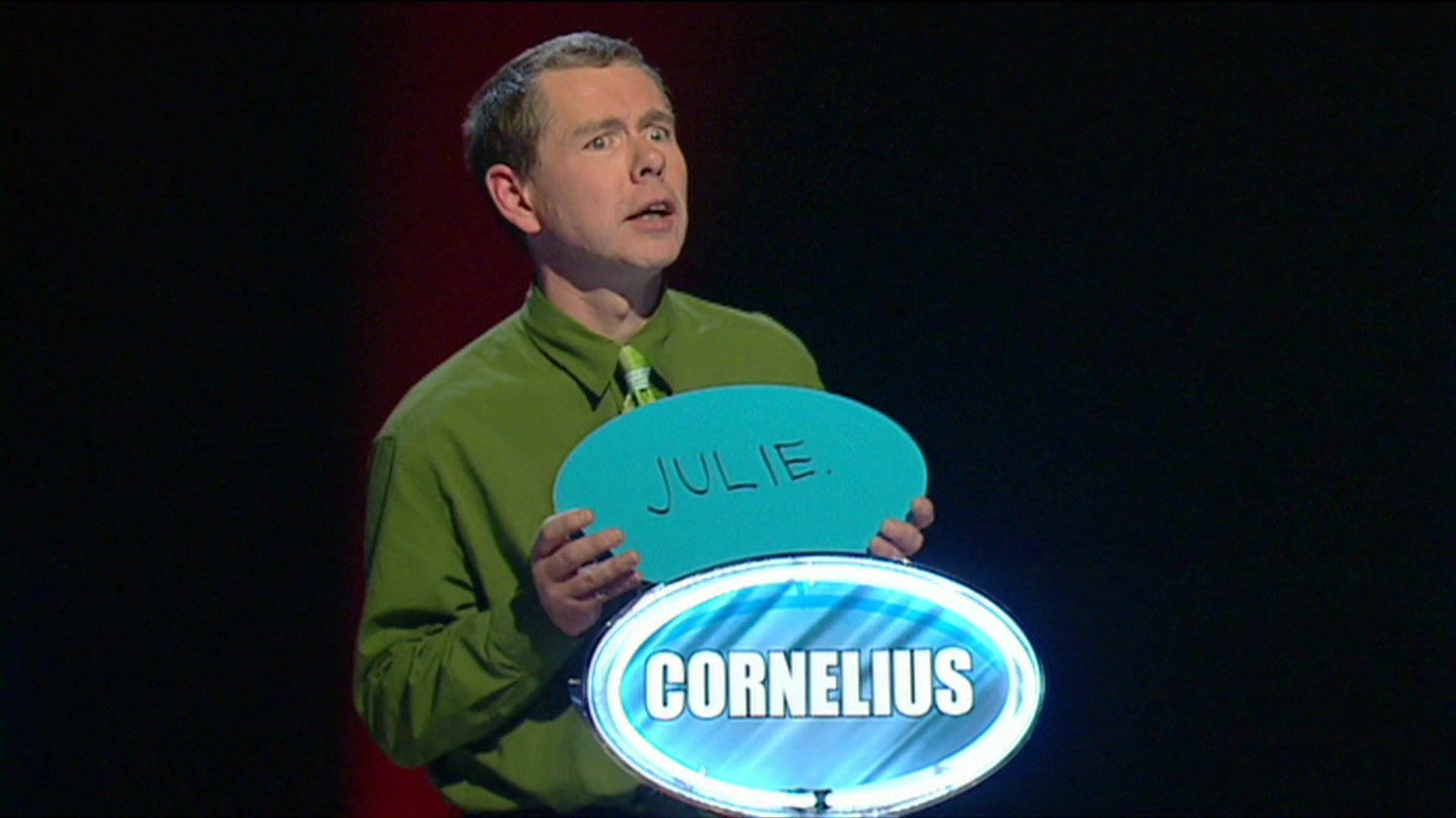 Cornelius O'Donovon wearing a green shirt and tie appearing on the Weakest Link with Anne Robinson. There is a sign in blue and white with his name on it and he is holding a sign with the name Julie on it.