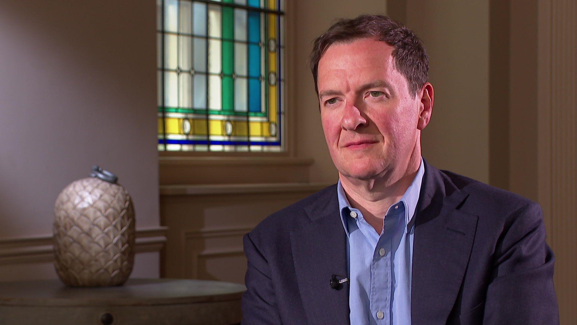 George Osborne is sitting down and starring towards the interviewer. He is wearing a navy suit and blue shirt.