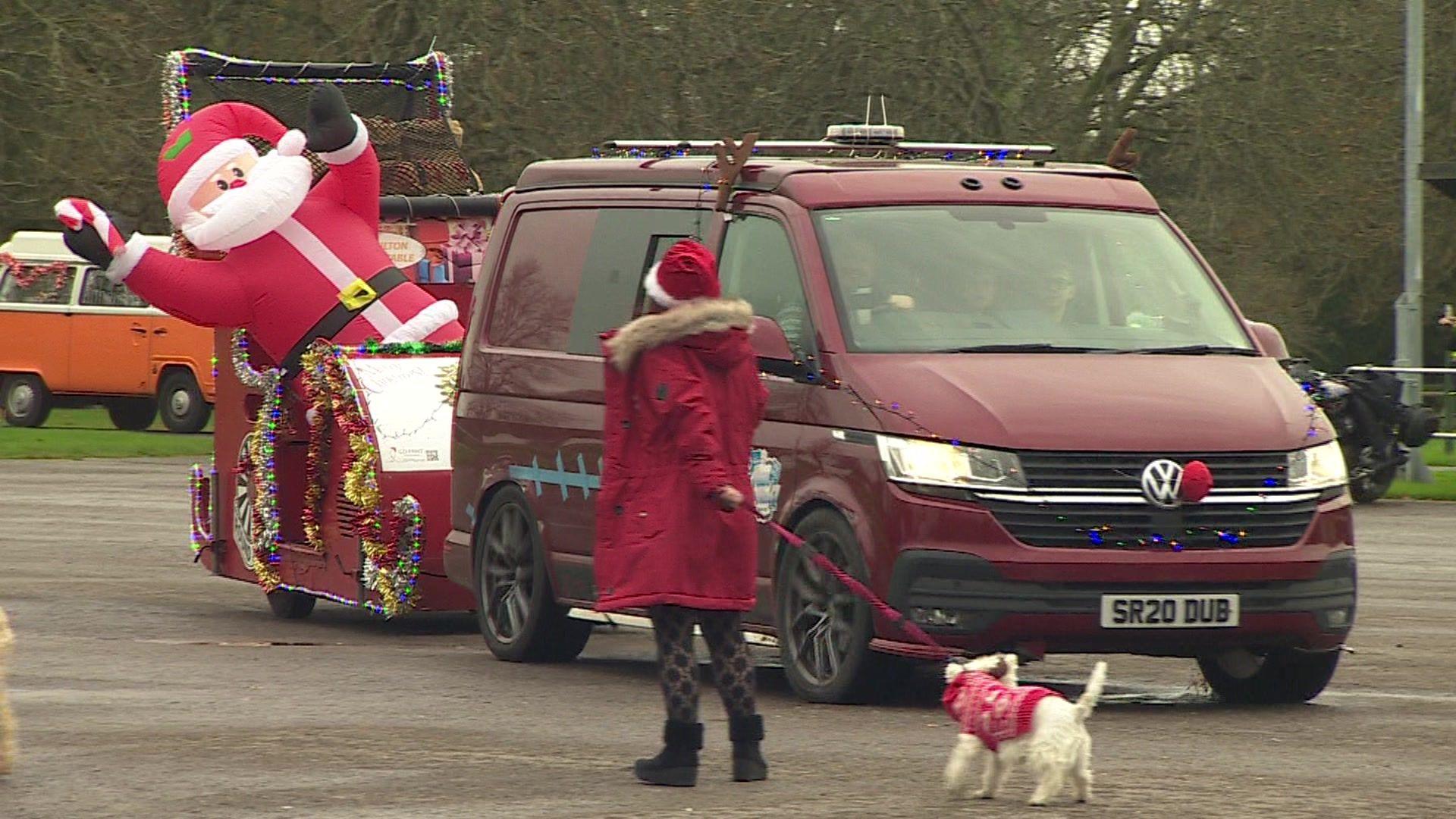 Red Volkswagen camper van driving in to Beaulieu Motor Museum. The van is towing a sleigh trailer with an inflatable Father Christmas on board. A woman in a red coat is walking a white dog in the foreground of the picture.