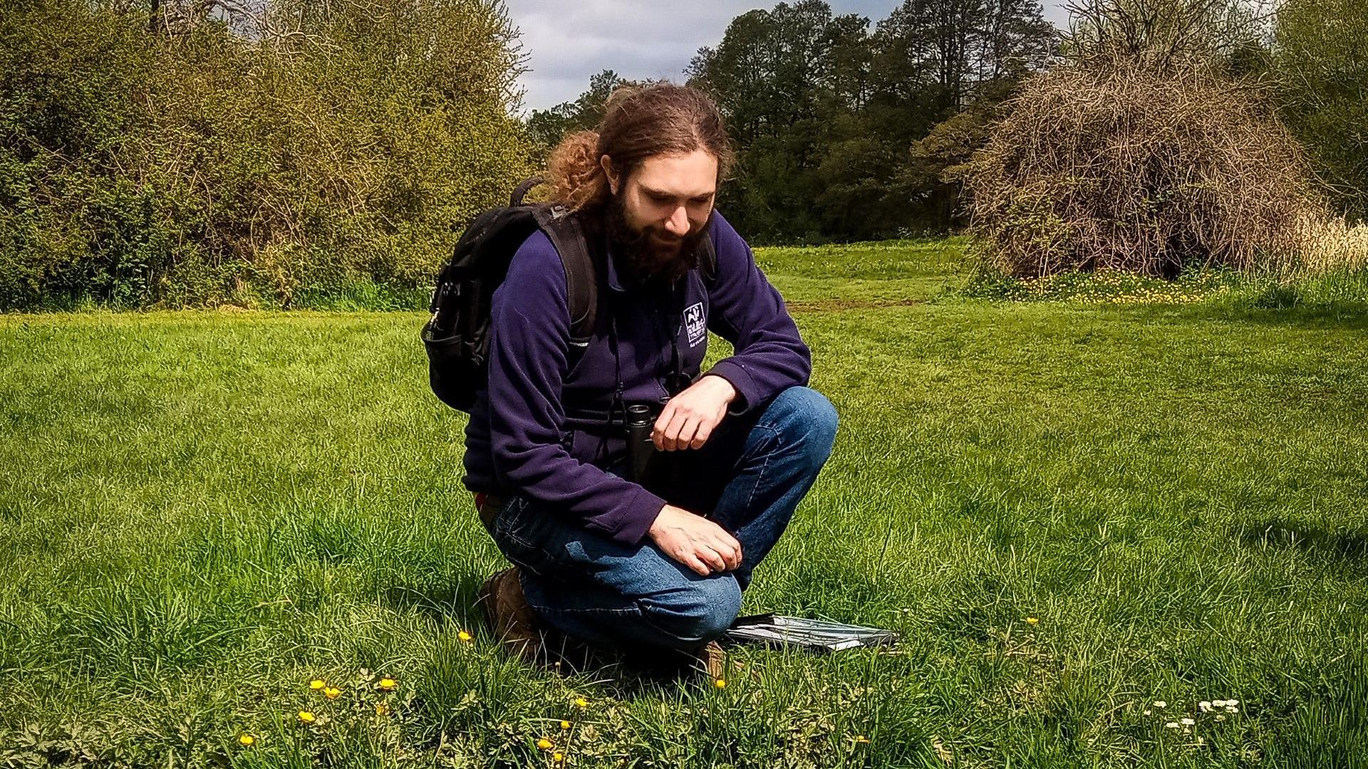 Alex Jones kneeling down in a field. It is a wooded area with lots of grass. There are trees behind him. He is bearded with tied-back long hair and wearing outdoor clothing. 