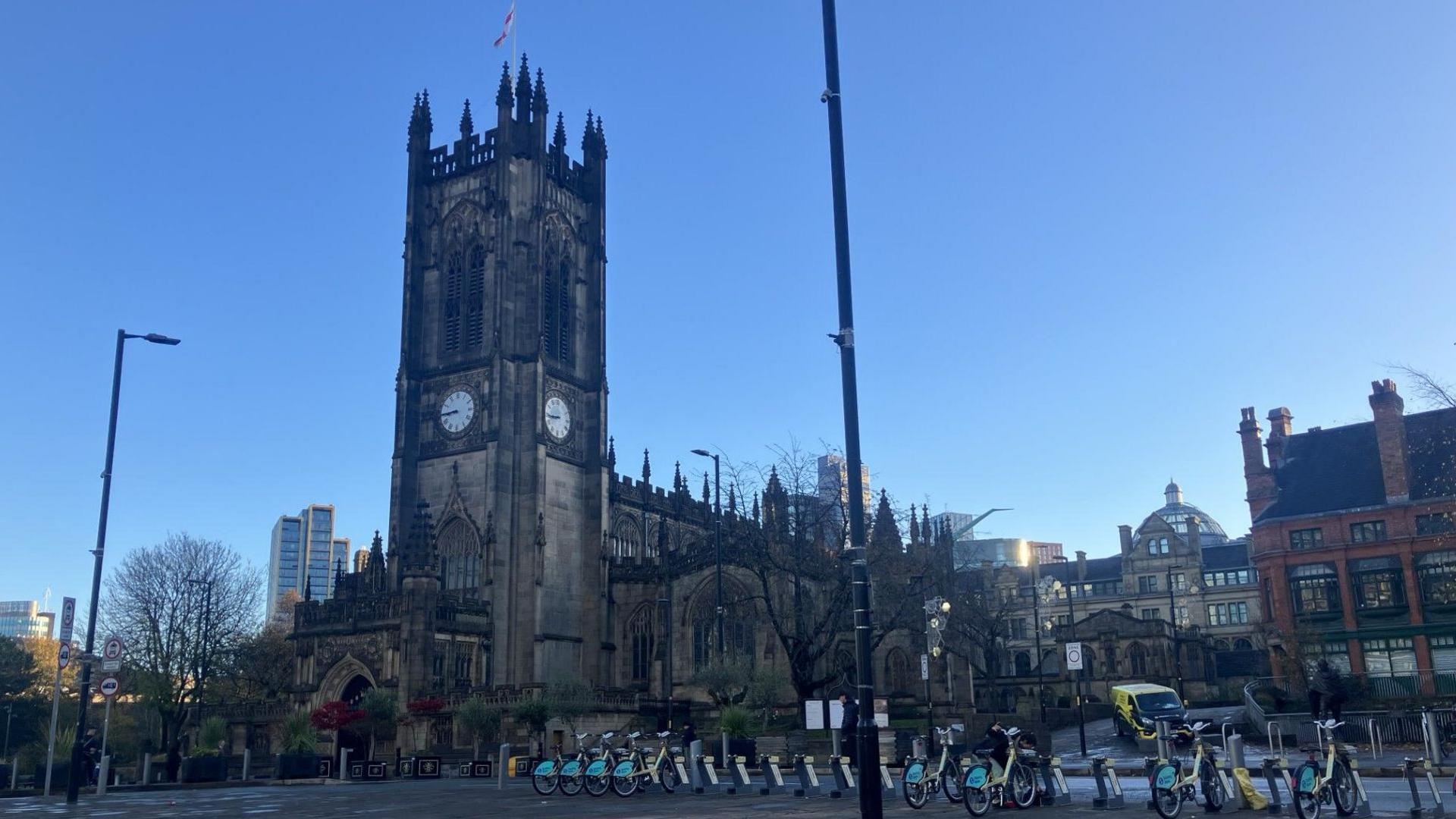 The photo shows a large Gothic-style cathedral with tall, pointed spires and intricate stonework. The building has a prominent clock face on its tower, and a flag is flying at the top. The cathedral is surrounded by a paved square with rows of rental bicycles neatly lined up in docking stations. In the background, modern high-rise buildings contrast with the historic architecture. The sky is clear and bright blue, suggesting a sunny day.