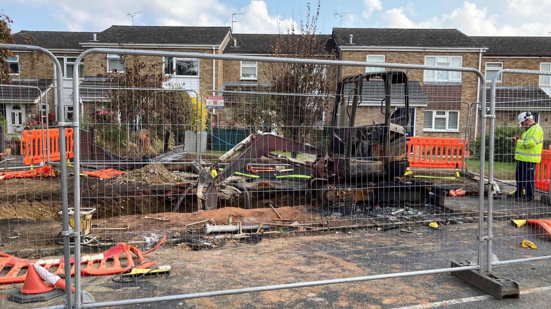 A burnt-out digger with its bucket in a trench, surrounded by temporary wire/metal fencing, with terrace housing in the background