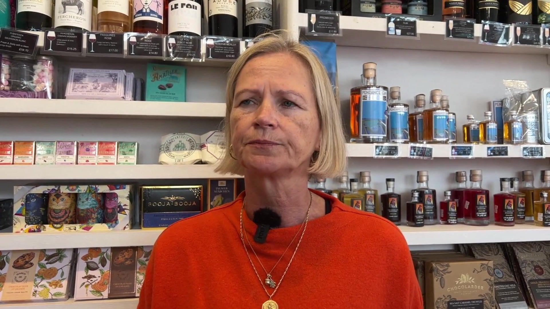 Sarah Shaw has been pictured in her store wearing an orange jumper, which she is wearing with two gold necklaces. She has blonde hair that stops just beneath her ears. Behind her are various white shelves with some of her products on them.