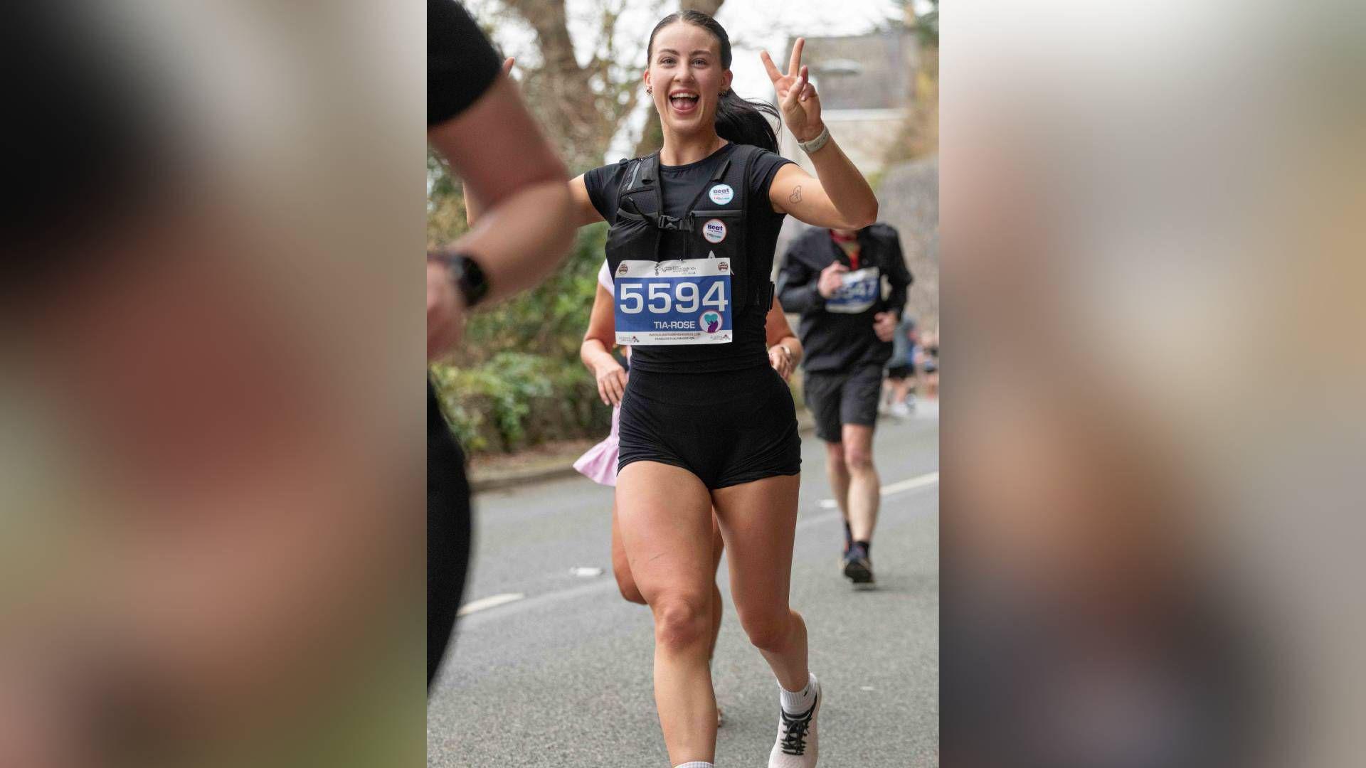 Picture of Tia Ainsworth running down a road in a parkrun, she smiles at the camera and holds both her hands up beside her head in two peace signs. She wears a running vest on top of black biker shorts and a black short sleeve t-shirt. She has a blue piece of paper attached to the front of her vest which reads: "5594 Tia Rose" in white font.  