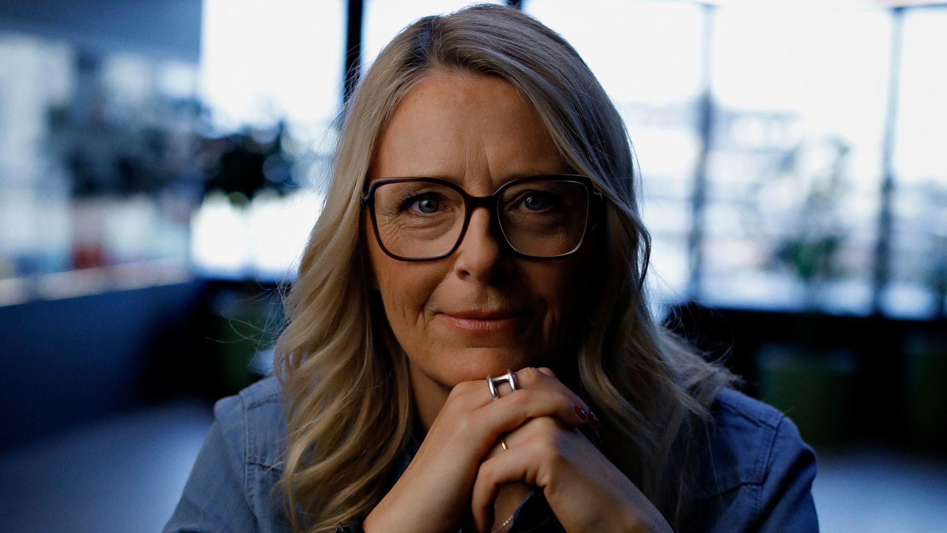 A woman with sleek blonde hair and black glasses poses for a portrait in an office setting. She's smiling with her chin resting on her hands.