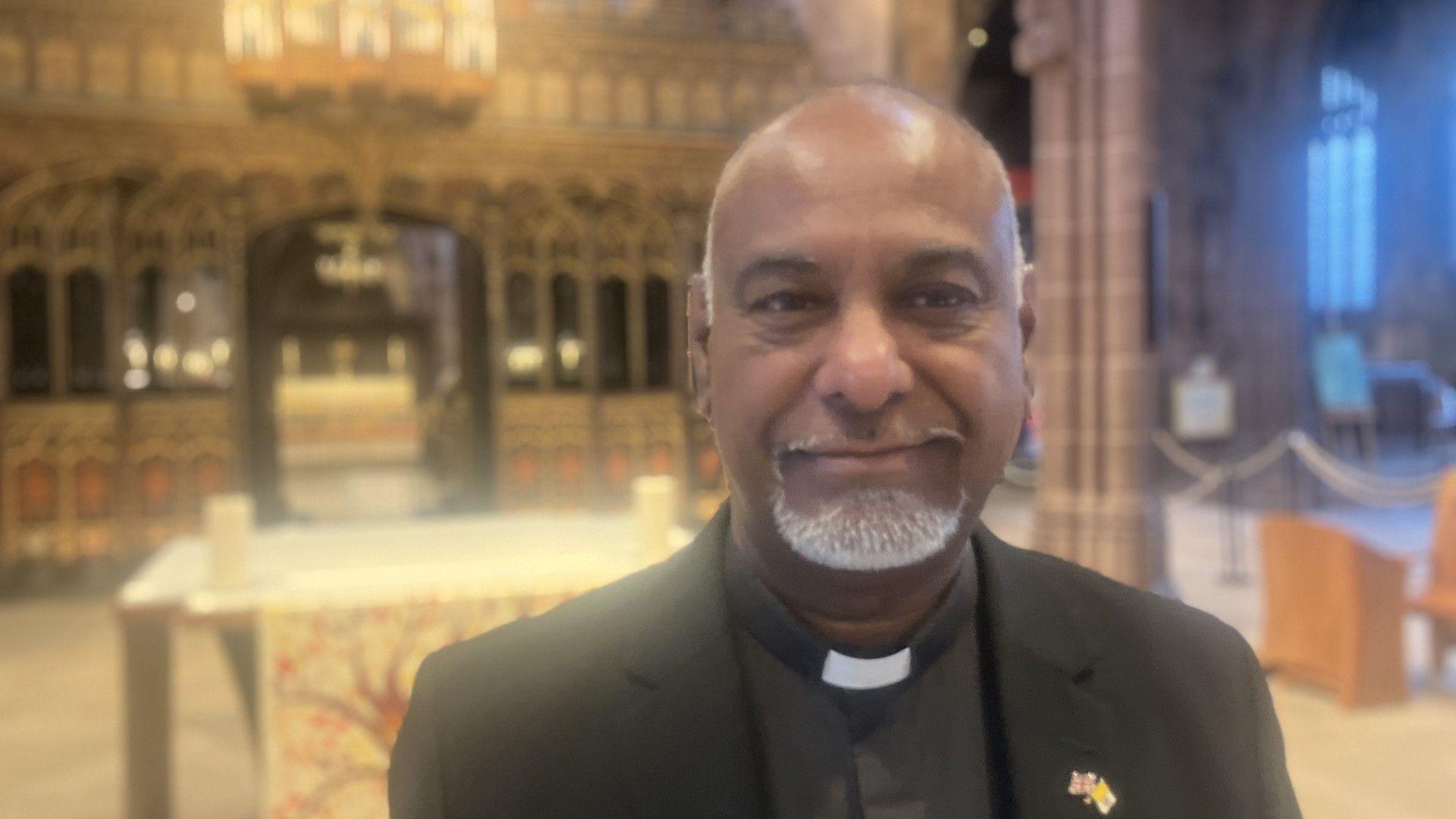 The person is indoors, in what appears to be the ornate interior of the cathedral. They are dressed in a black clerical suit with a white clerical collar, indicating a religious role. A small pin with a flag design is visible on the left lapel of the jacket. Behind them is an elaborate altar with gold detailing and decorative woodwork.