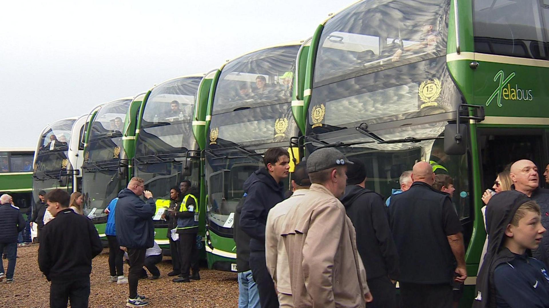 A row of green double decker buses with football supporters milling about around them.