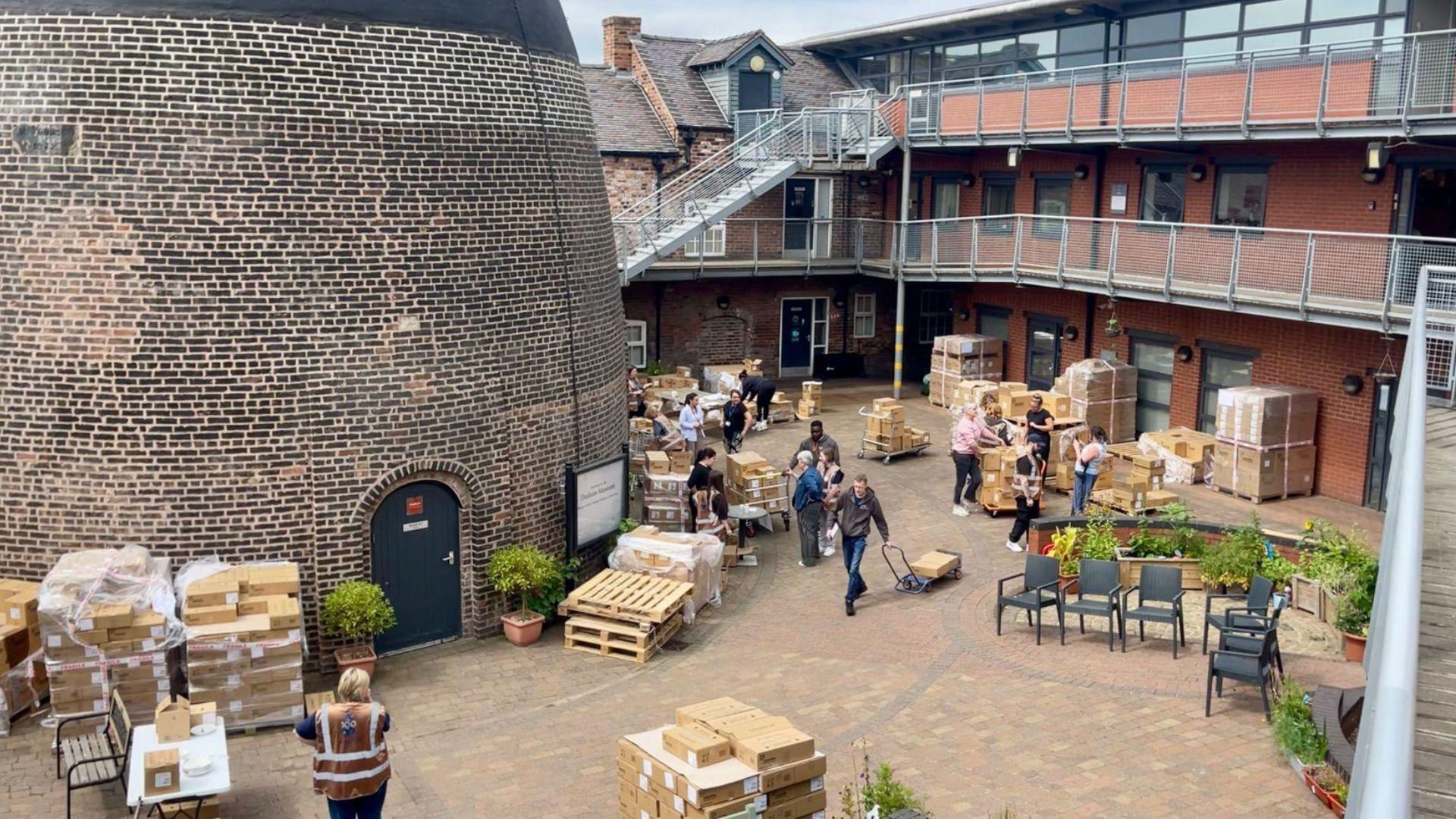 A huge brick bottle kiln is surrounded by pallets piled with cardboard boxes. Fifteen people, some in gold tabards, can be seen standing by the boxes. One man is dragging a trolley with a cardboard box on. There are also patio chairs and garden plants. There are tree storeys of offices in the background with steel railings and steps leading to them.