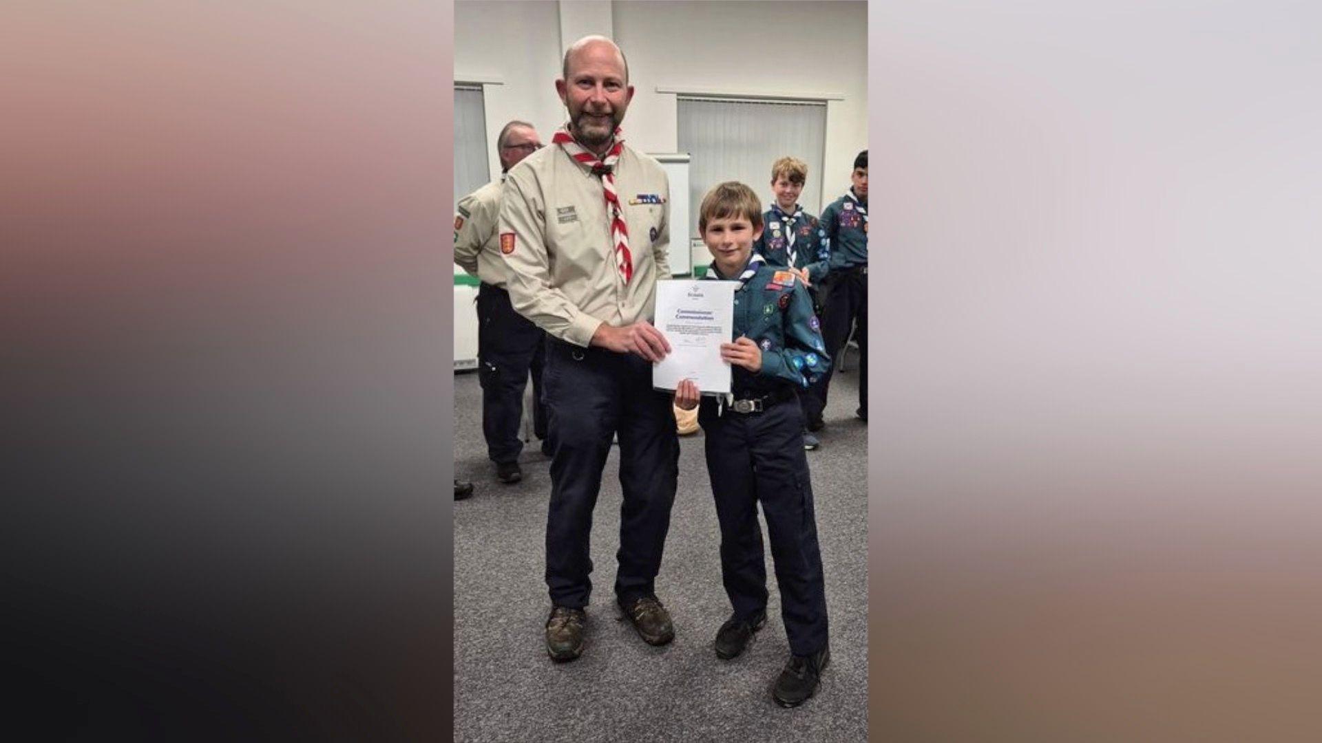 Glen and Charlie smile at the camera as they hold a certificate between them. They're both wearing Scouts uniforms and there a few Scouts behind them. Glen has a cream coloured shirt with a red and white neckerchief. Glen has a stubbly beard and a bald head.