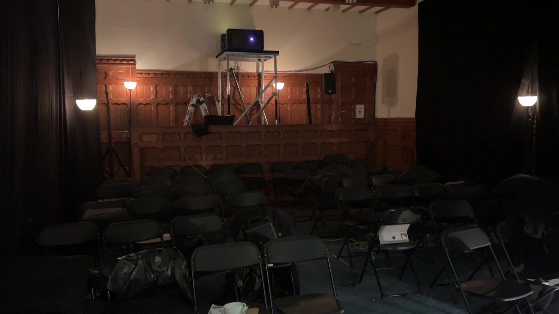 A small courtroom with a large projector behind a wooden divider. There are rows of black chairs in front of it.