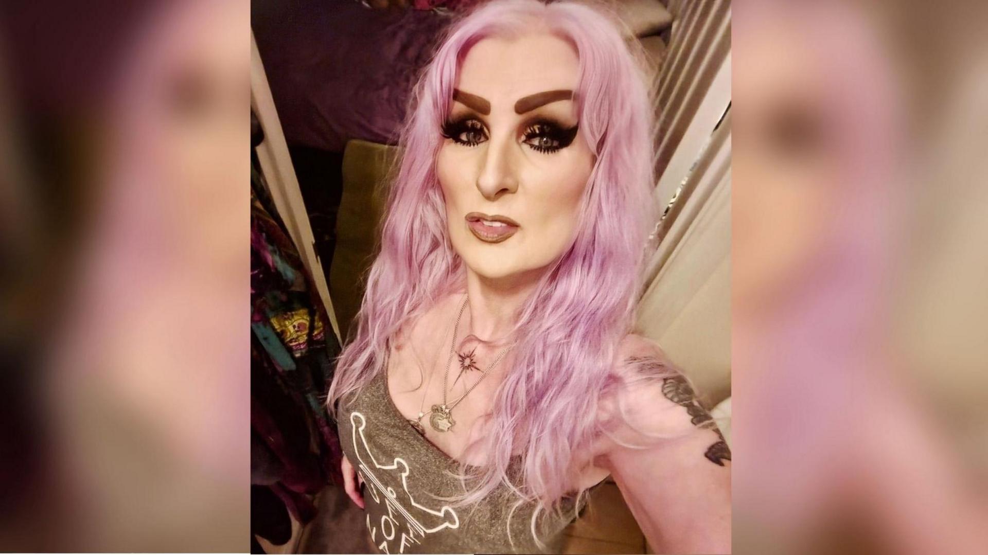 Wendy has long pink hair, heavy eye make-up and tattoos visible on her left shoulder. She is wearing a vest top with a white logo on the front and a necklace. She is taking a selfie and partially smiling at the camera.
