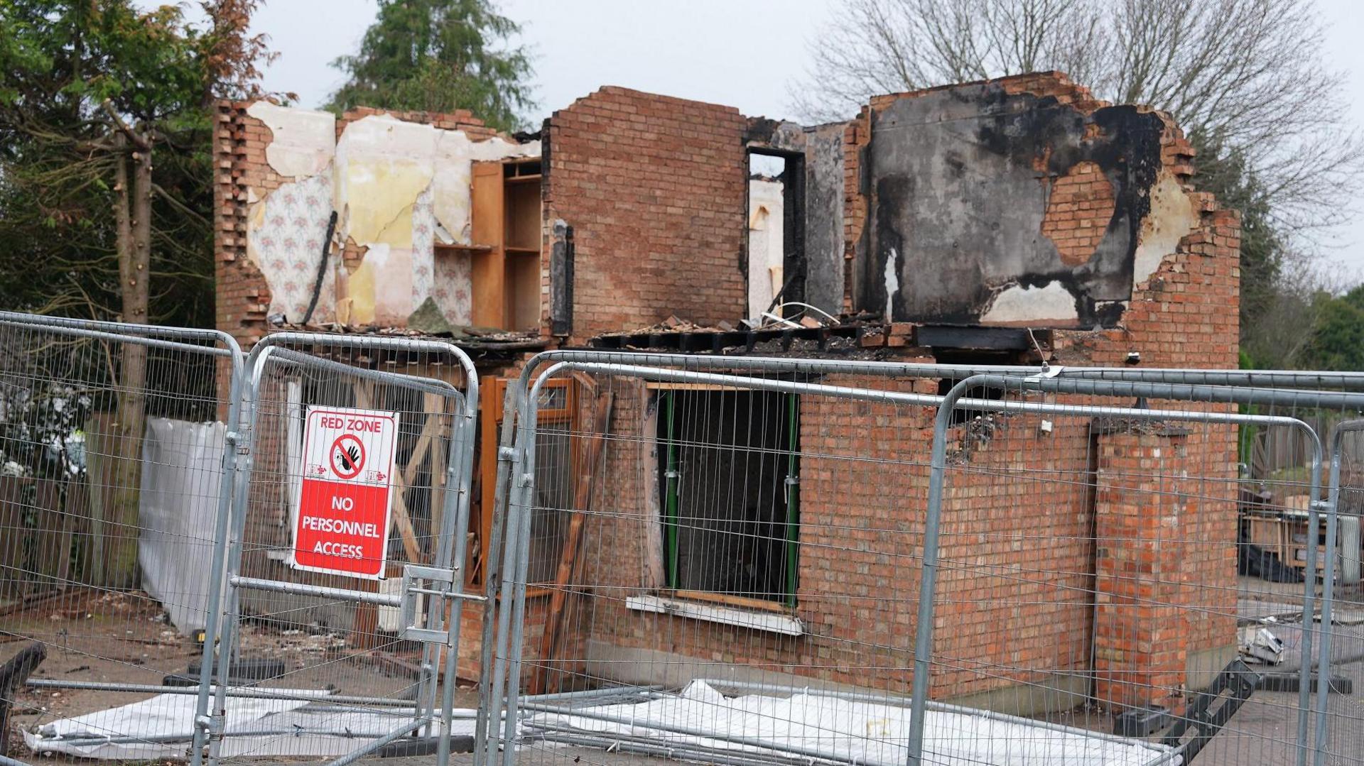 Metal barriers  in the front of the damaged house, with a sign attached saying "Red Zone - No Personnel Access". the brick house has the top floor mostly destroyed, with blackened wallpaper on the back and side walls. The window downstairs is missing and there are white tarpaulins on the floor. 