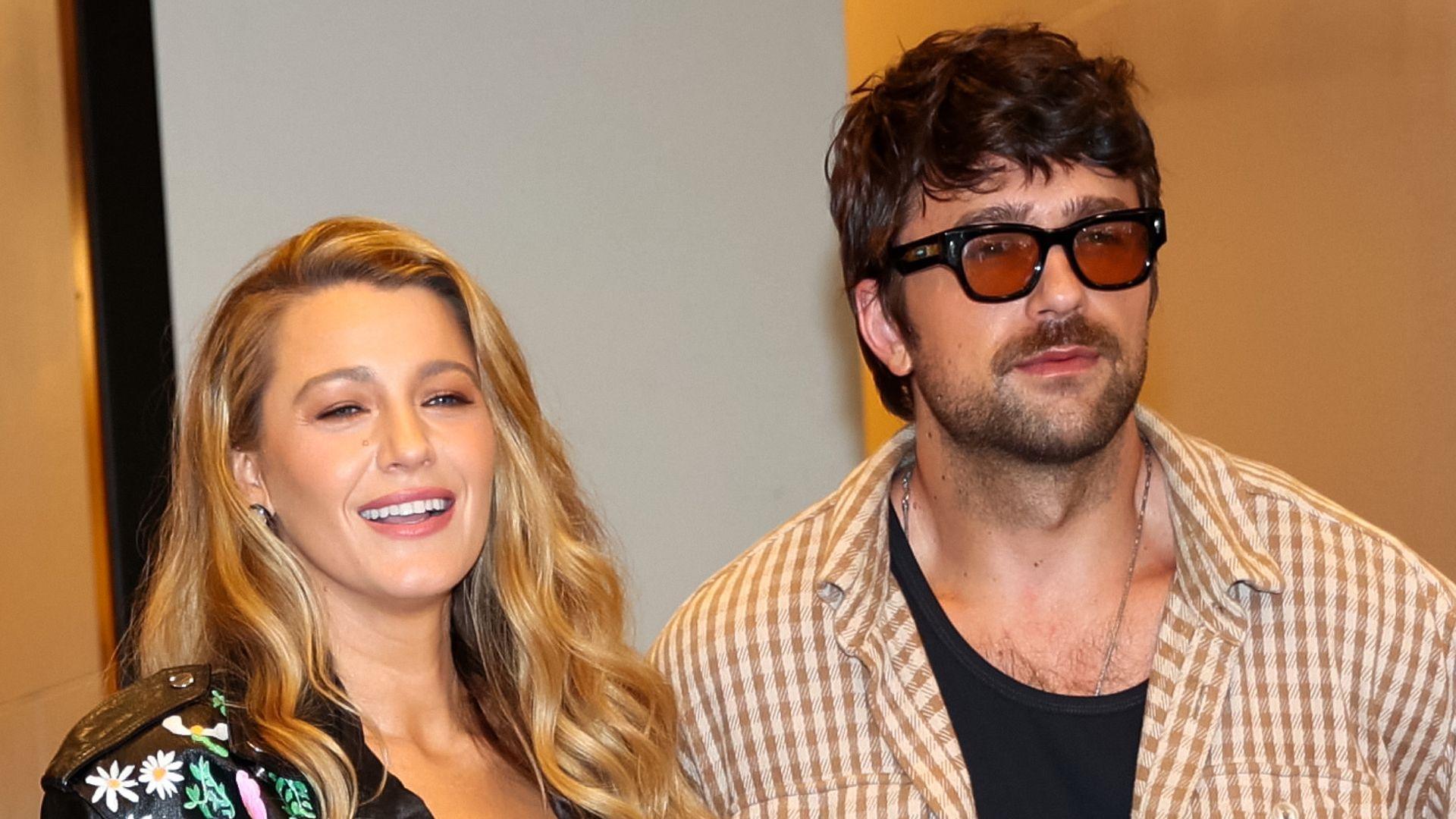 Blake Lively and Brandon Sklenar pictured in New York in August. Blake is a woman in her 30s with long curled blonde hair. She wears a painted leather jacket and smiles. Brandon, a 34-year-old man, has shaggy brown hair and a short stubbled beard. She wears brown tinted sunglasses and a brown gingham shirt over a black vest.