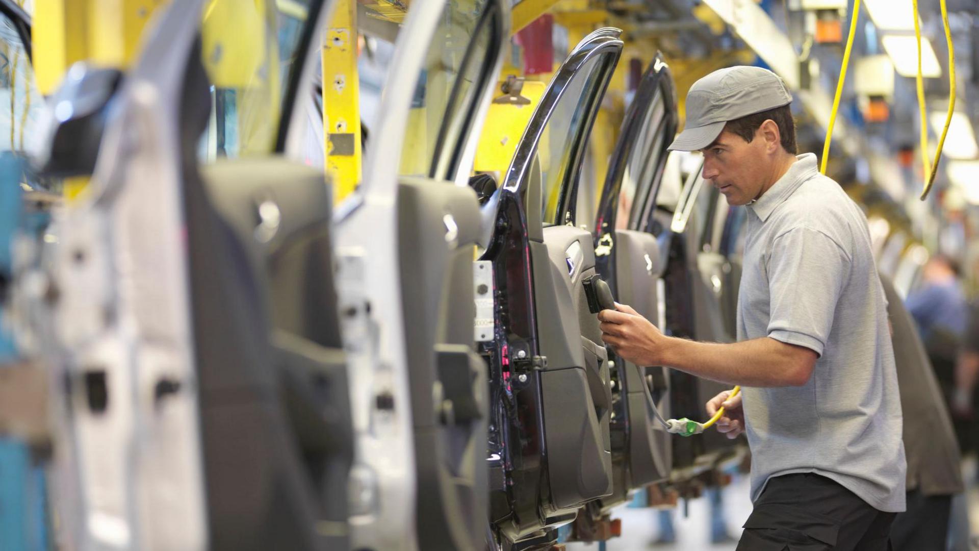 A worker wearing a grey polo shirt and baseball cap inspects car doors on a factory production line.