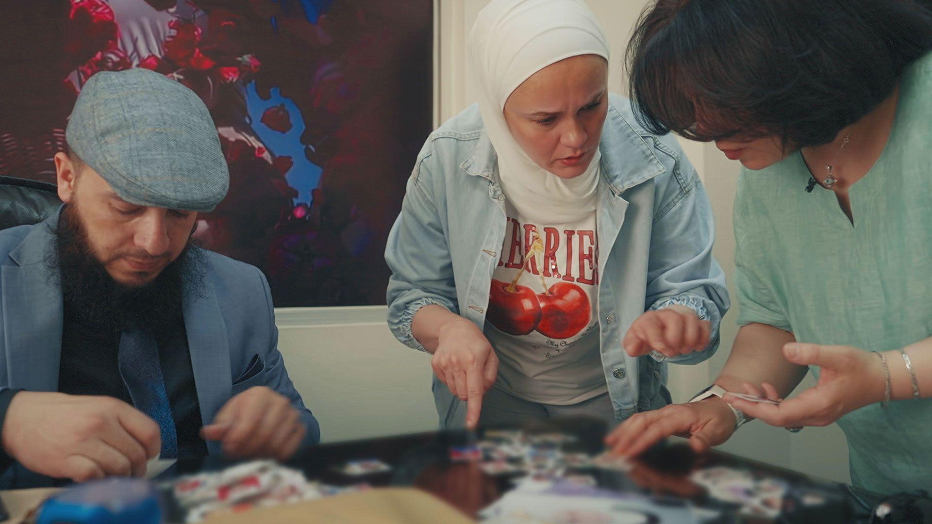 The manager of the children's shelter is wearing a blue jacket and blue tweed cap. Reem is wearing a blue shirt open over a t-shirt with cherries on it and has her hair covered by a white headscarf. They are all looking through photos of children spread out on a desk.