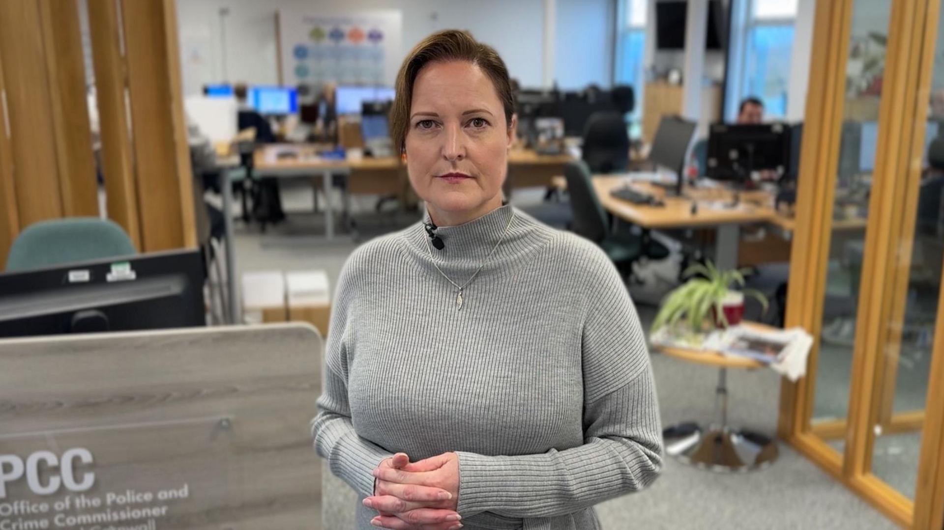 Alison Hernandez with a stern expression wearing a grey jumper, standing in an office