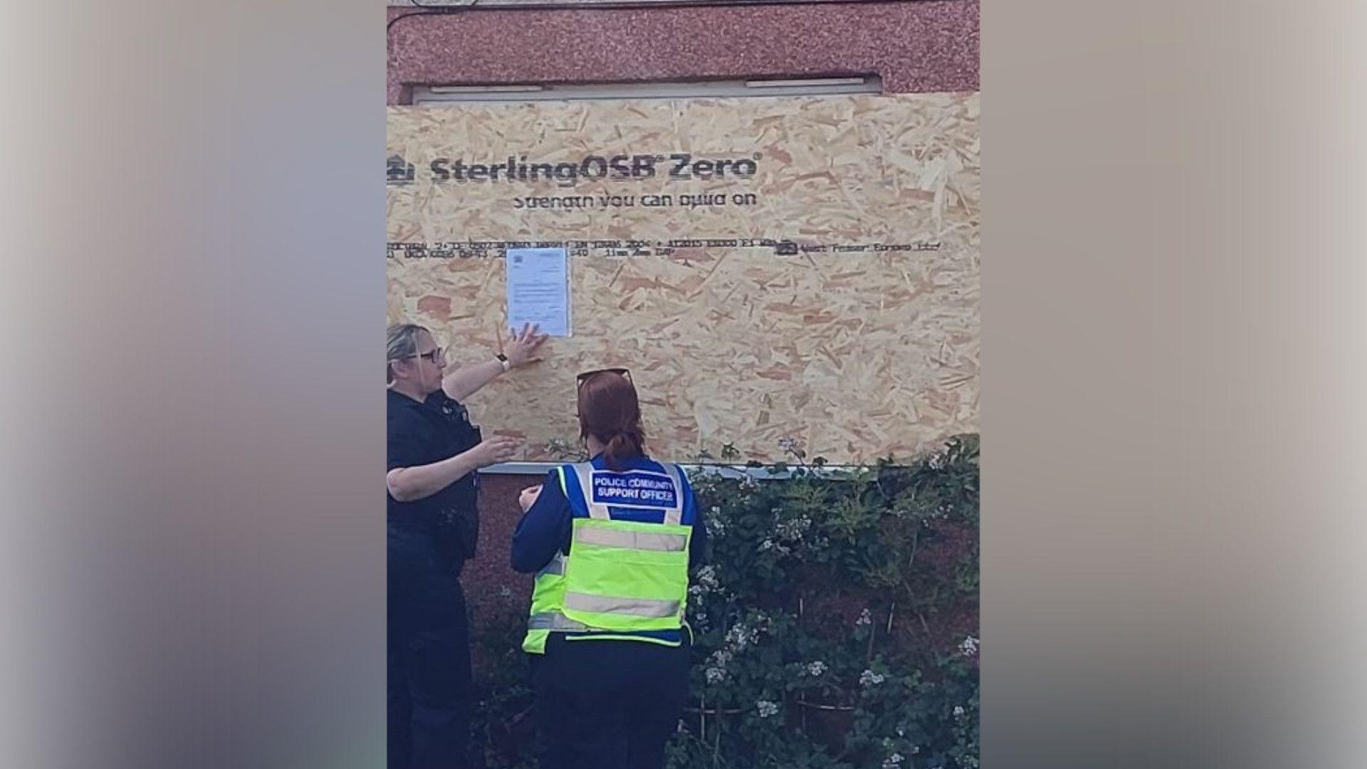 A female police officer in a black shirt is attaching a piece of paper to a wooden board fixed over the window of a house. The board says "Sterling OSB Zero". A female police officer with long brown hair and wearing a yellow hi-vis tabard is looking on. The red render of the house is visible above and below the board. There is a hedge in front of the board.