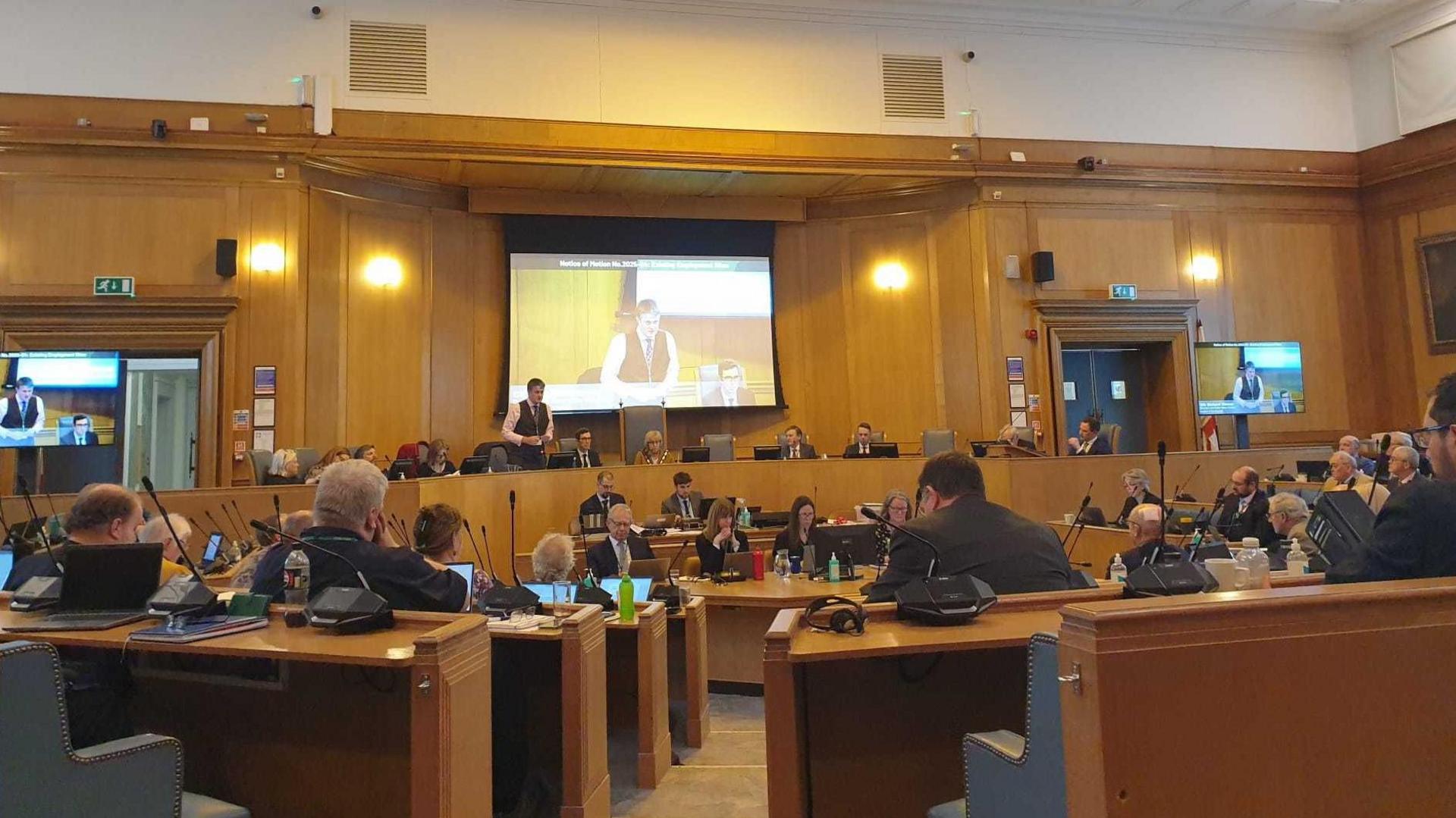 A council chamber, with lots of wooden benches with microphones and wooden panelling. People are dressed smartly, looking to the front where there are larger benches with chairs facing the rest of the room and a big screen behind.