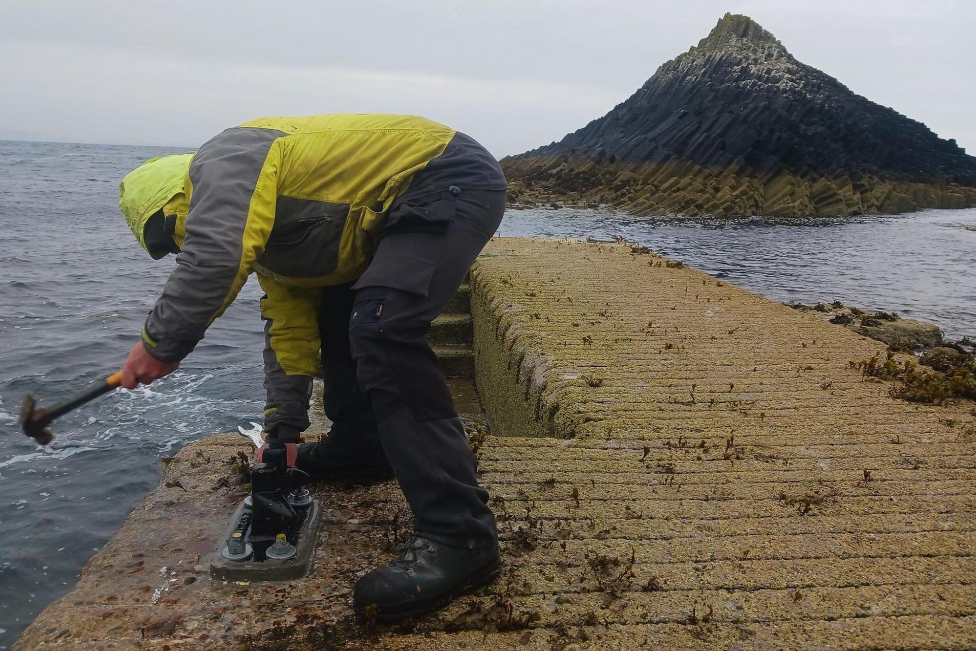 A worker wearing a yellow jacket with the hood up, work trousers and sturdy boots is bent over as he used a hammer at the jetty. It is a wet and windy day and there is a rocky outcrop in the distance.