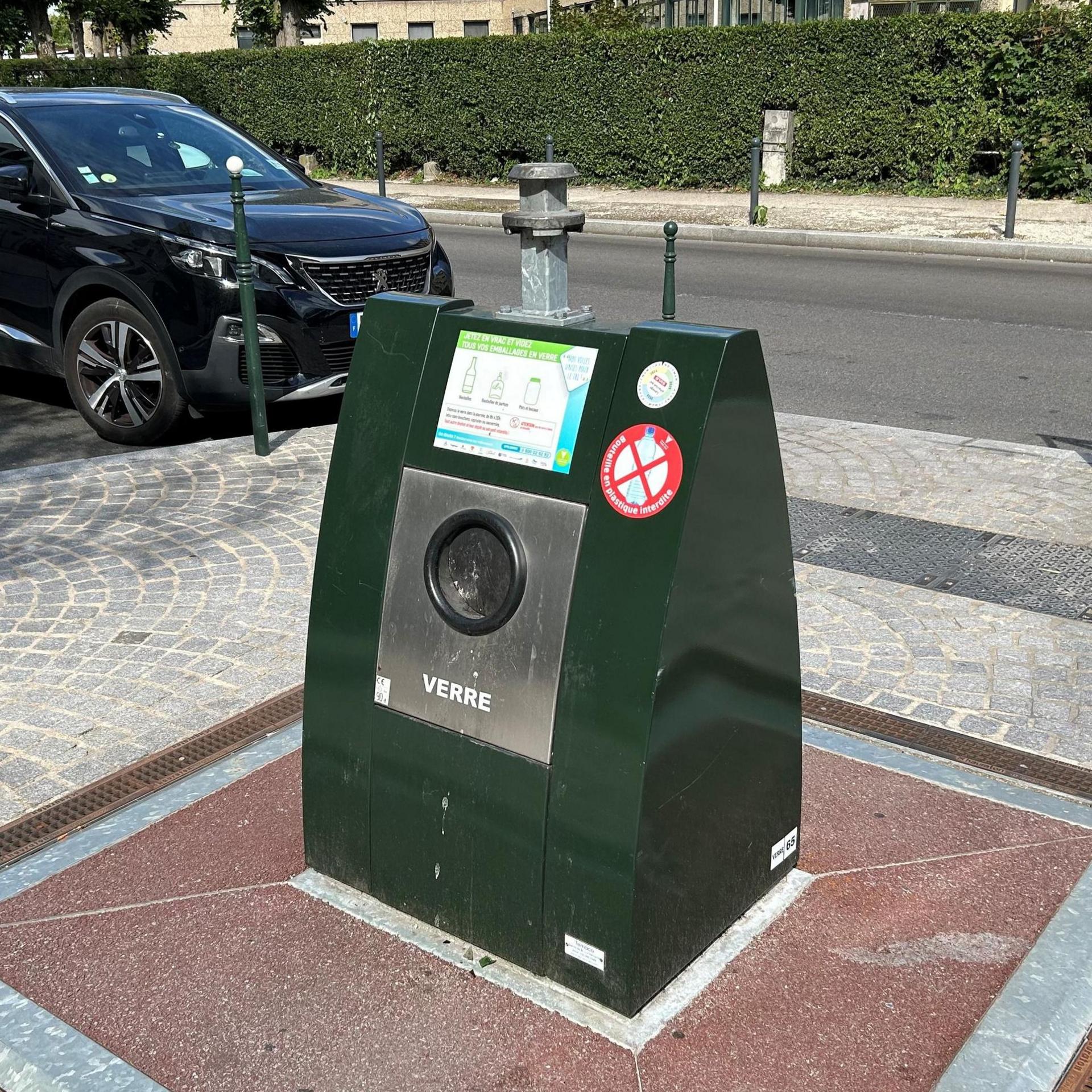 A large green subterranean communal bin installed in a paved area on a residential street. The bin has a circular opening on the front and a sign with disposal instructions. Cars are parked along the road in the background, with a hedge and apartment building behind.