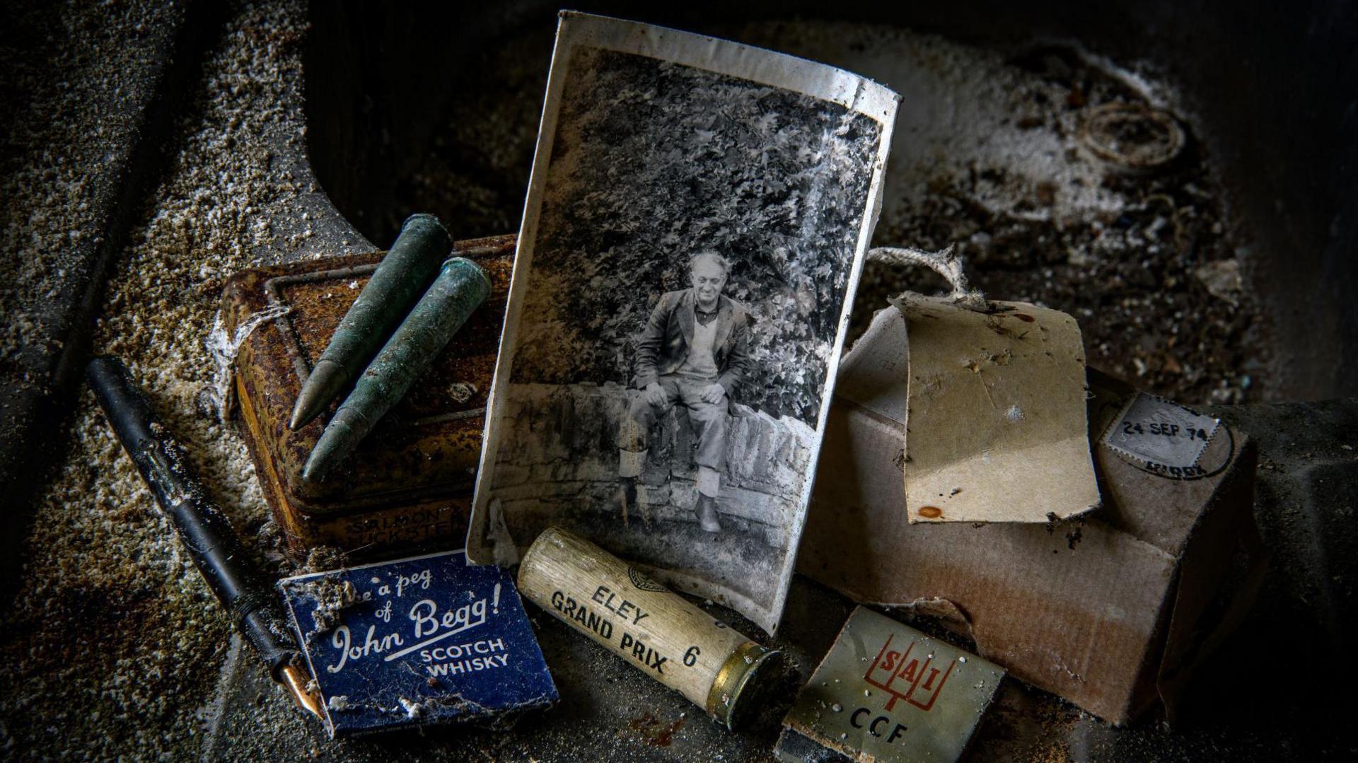 There is a collection of objects on a dusty floor. They include an old photograph of a man sitting on a wall, matchbooks, gun cartridges, a fountain pen and rusty bullets.