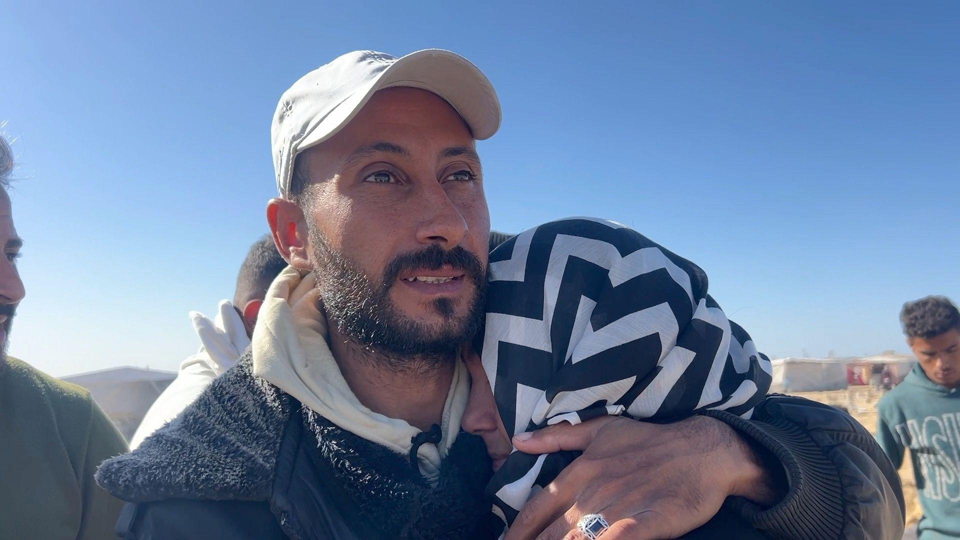 A man holds a woman wearing a black and white headscarf against him