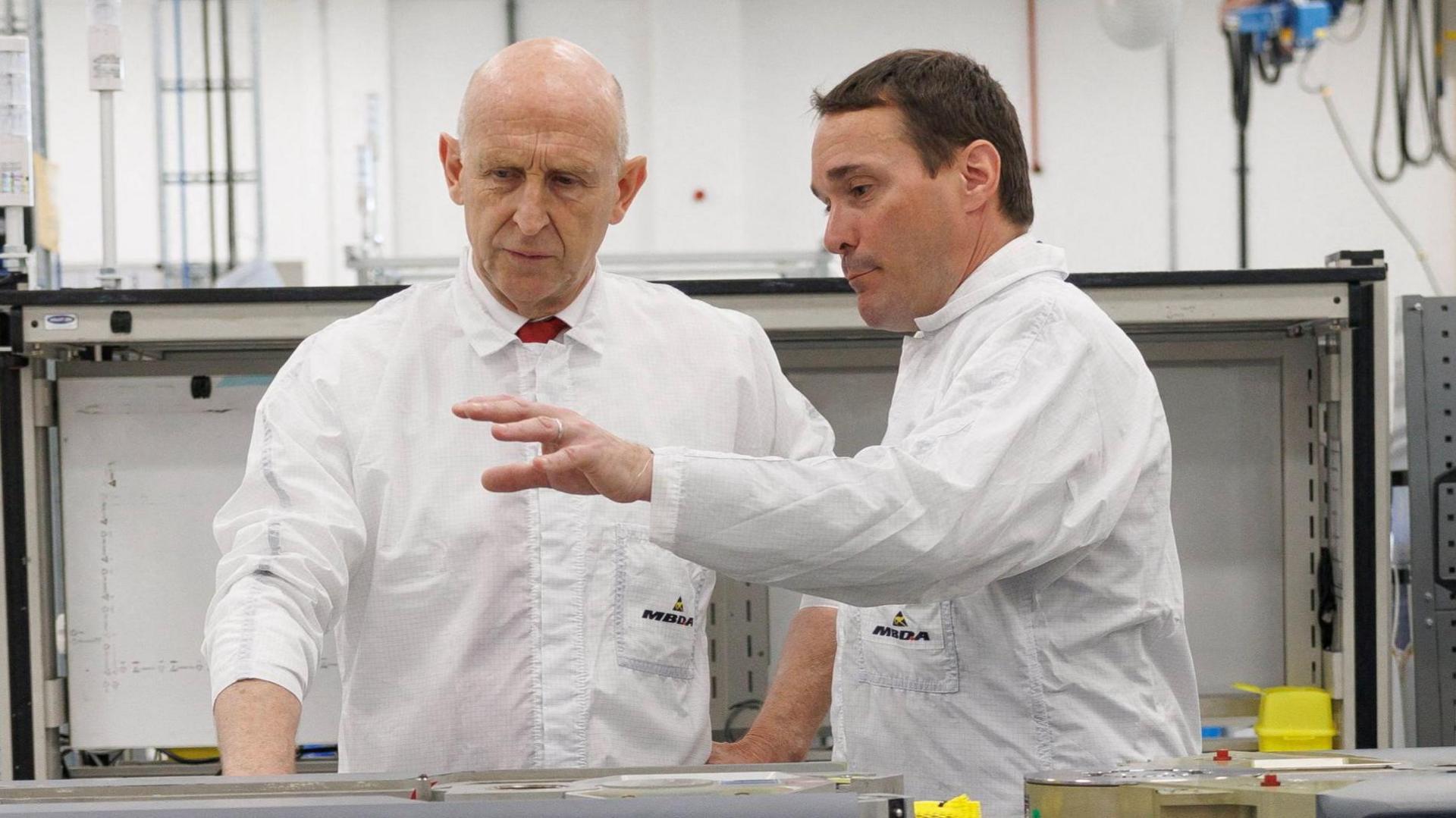 Defence Minister John Healey speaks to a man on an assembly line at a factory in Stevenage. Healey is bald and has blue eyes.