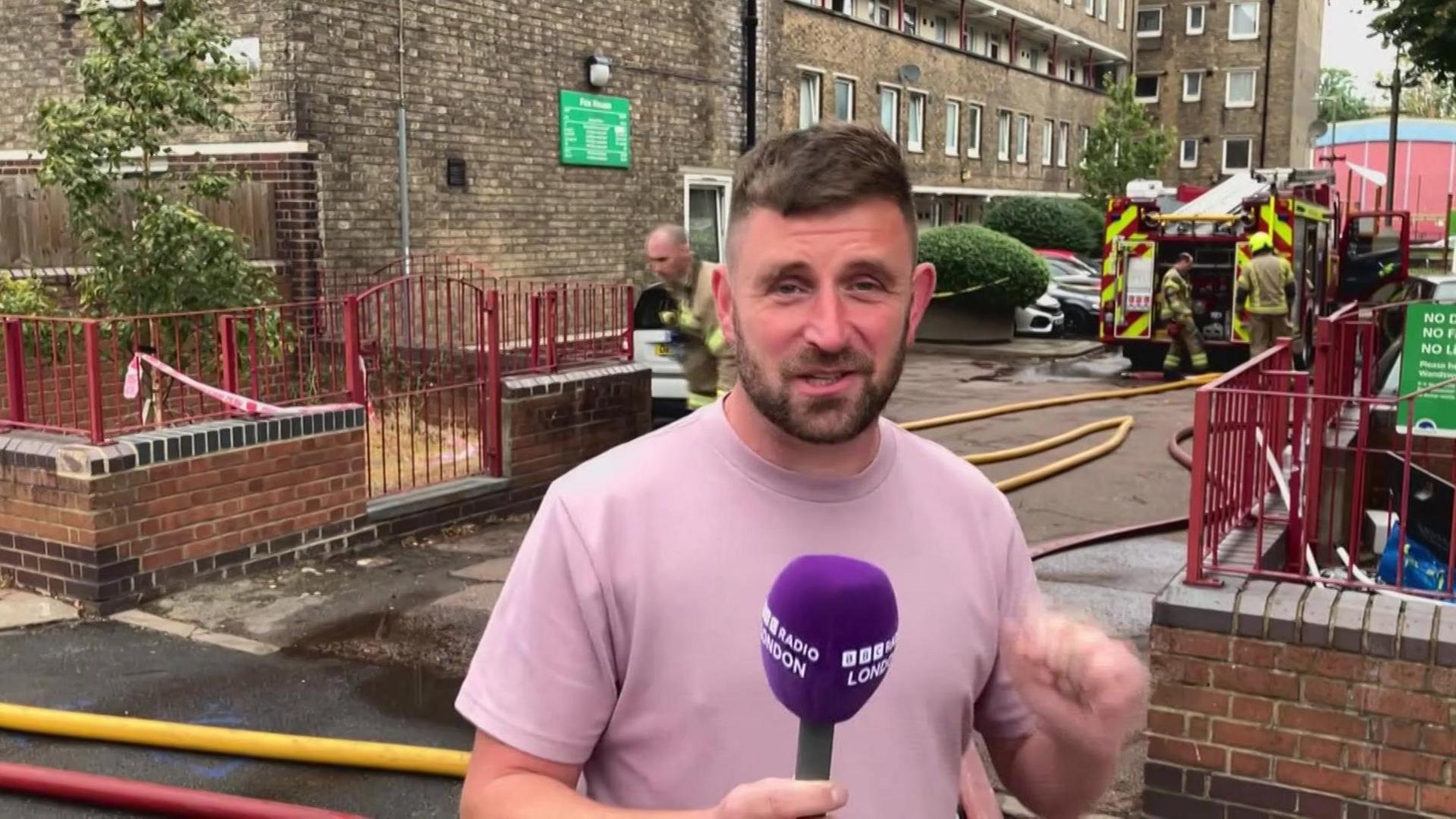 A man in a pink t shirt with short brown hair and a short brown beard stands in front of a block of flats. Fire brigade hoses and a fire truck as well as some firefighters are in the background.