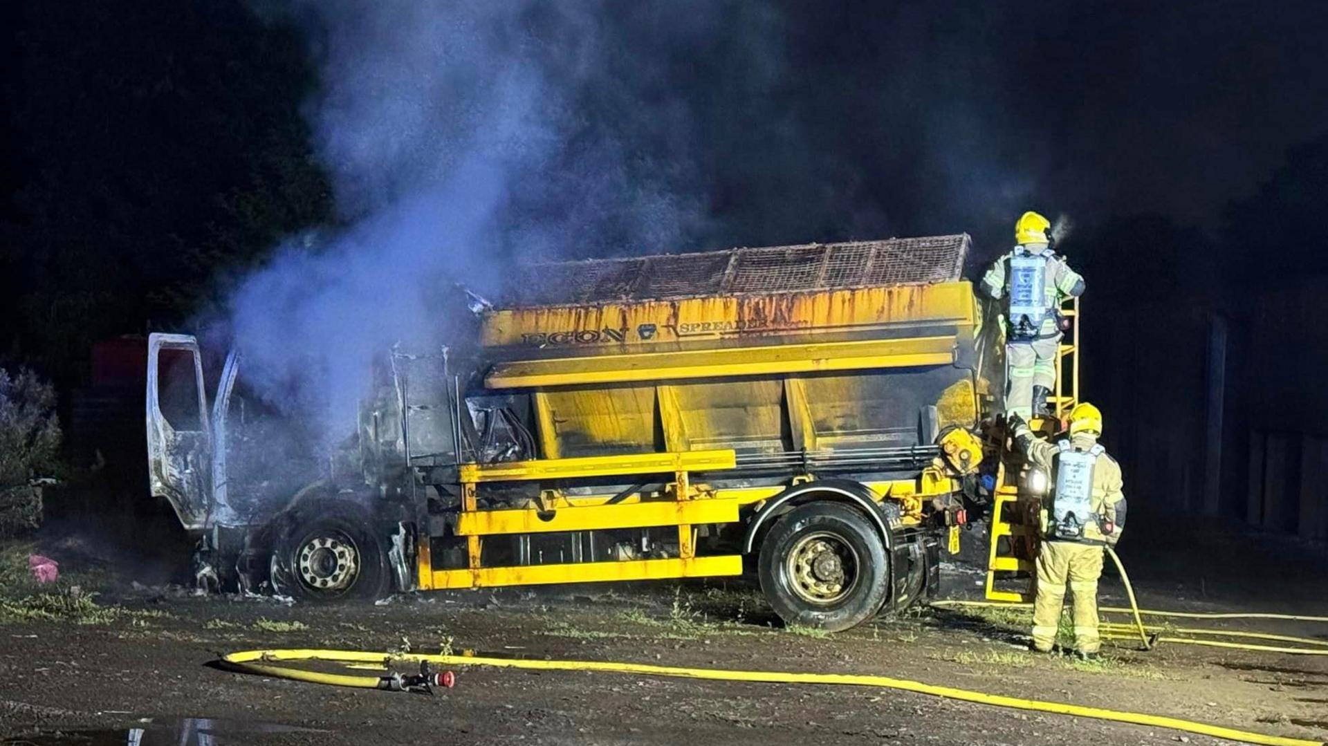 Smoke surrounds a yellow gritting lorry under dark skies, the front of the vehicle is burnt out.

Two firefighters are at the back of the vehicle with hoses