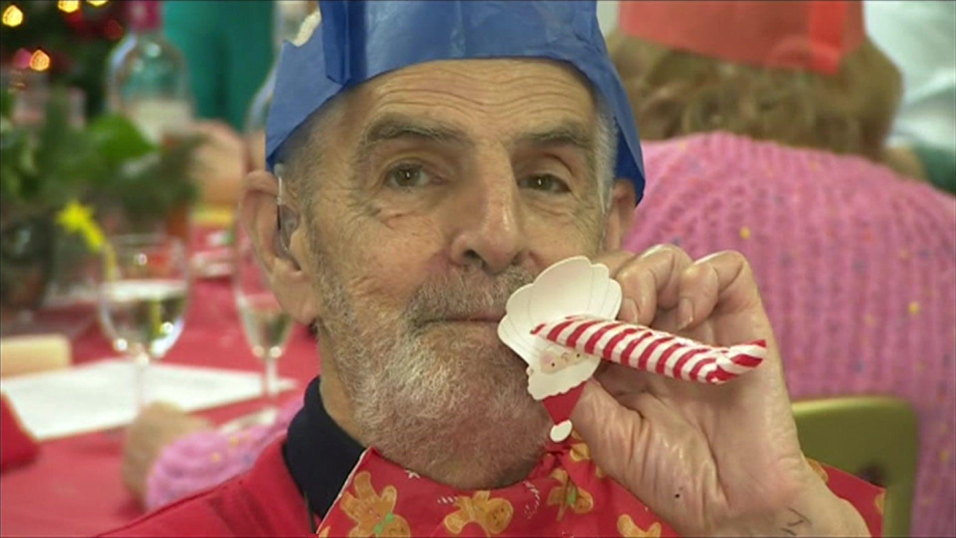 A man is blowing a party blower that is red and white. He is wearing a blue Christmas cracker hat and is wearing a red jumper.