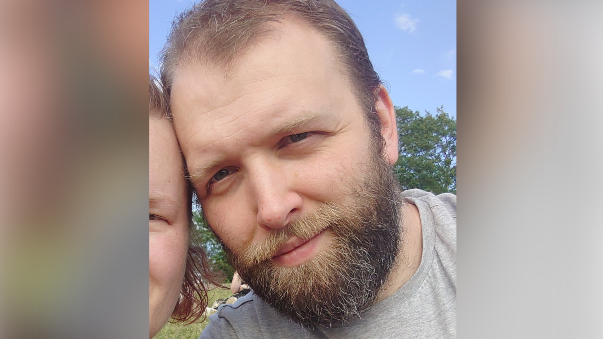 A man leans his head against a woman (who is just in the picture). He has a beard and is smiling.