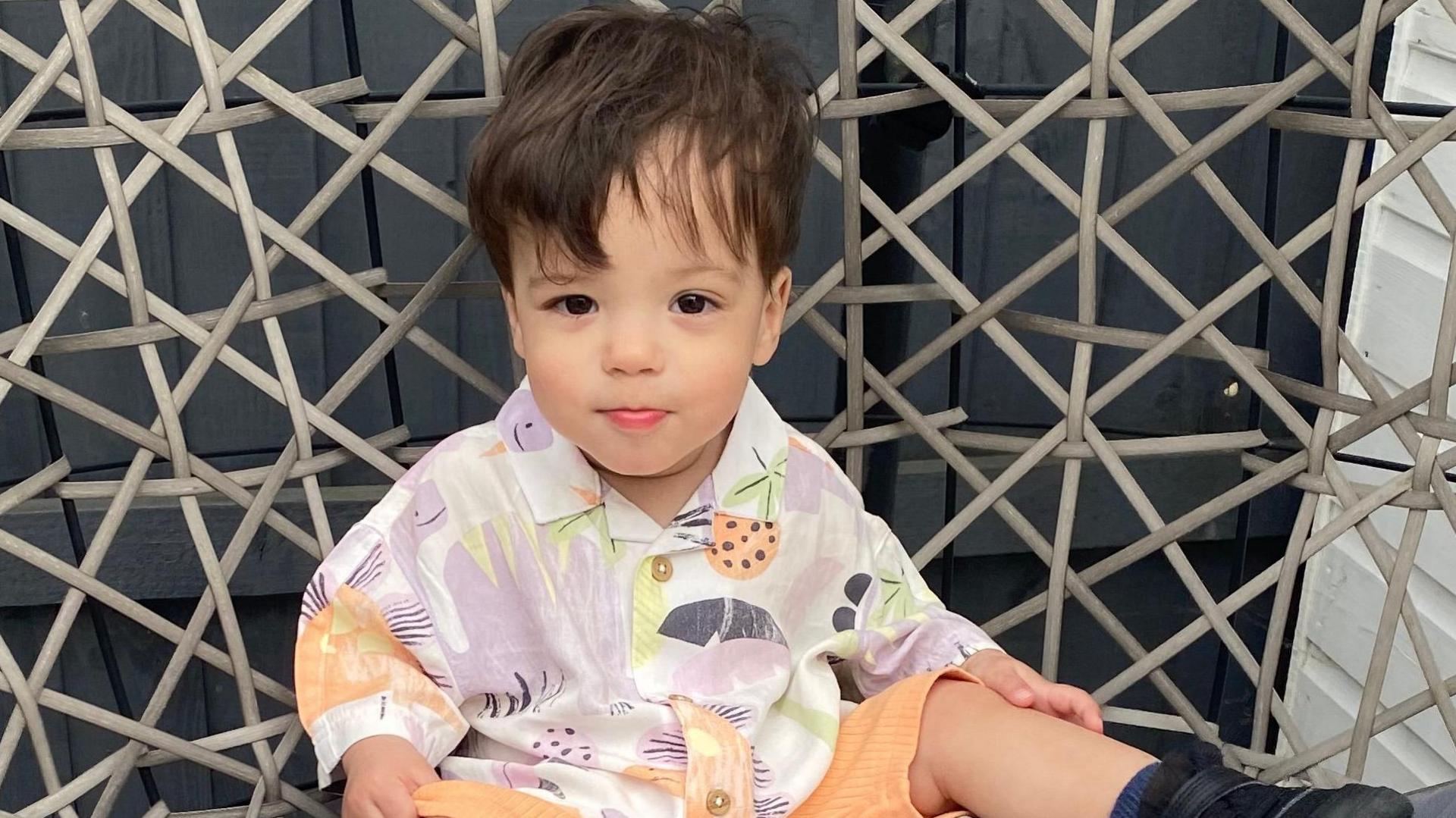 A baby sat on a wicker chair, looking at the camera