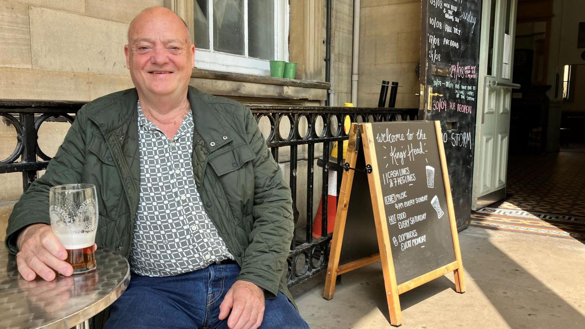 A smiling man sat outside a pub with a pint of beer. He is wearing a green jacket, a green and white patterned shirt and blue jeans.