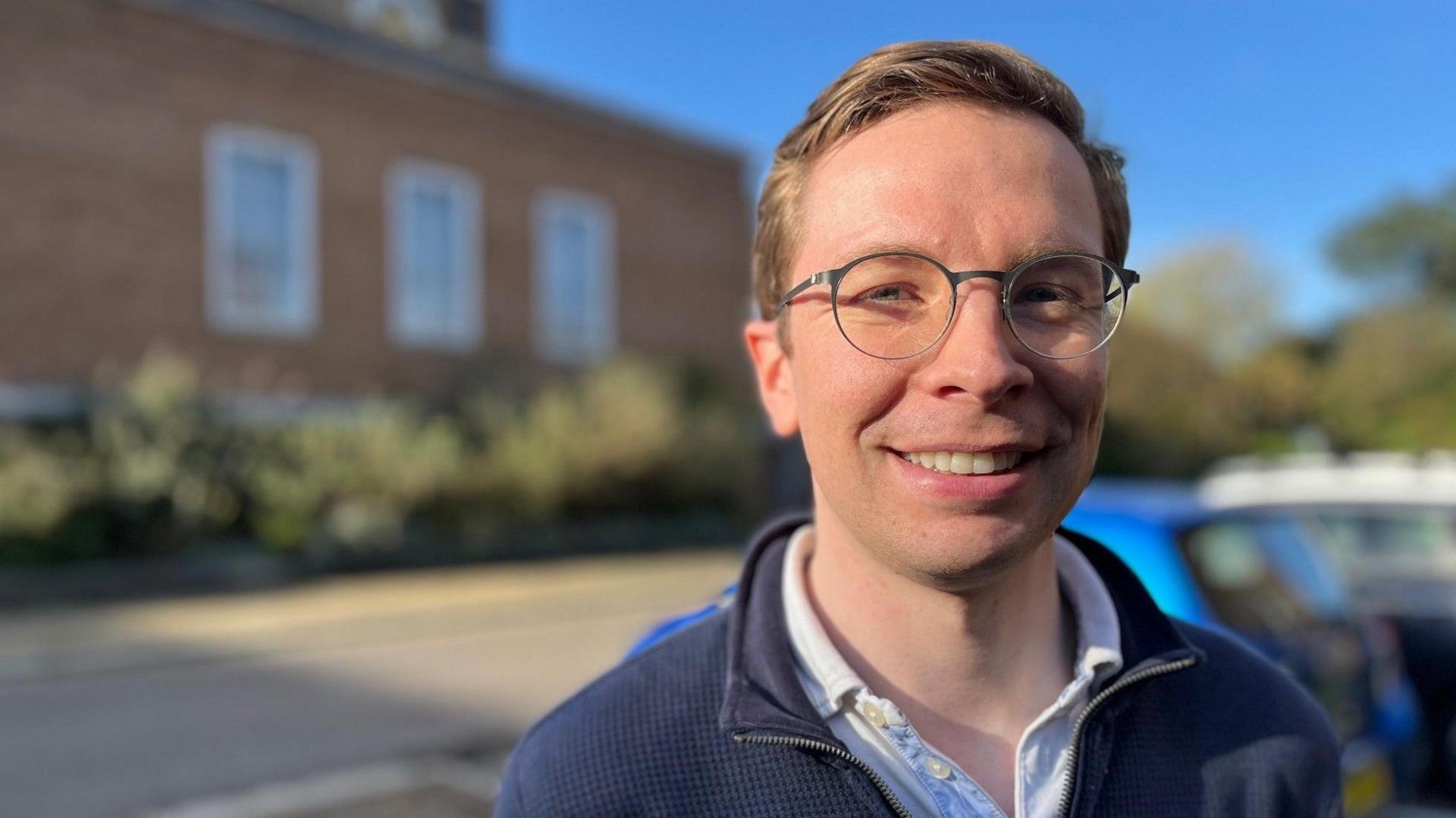 Karl Wiseman, wearing glasses, a light shirt and a blue zip-neck jumper against a blurred background