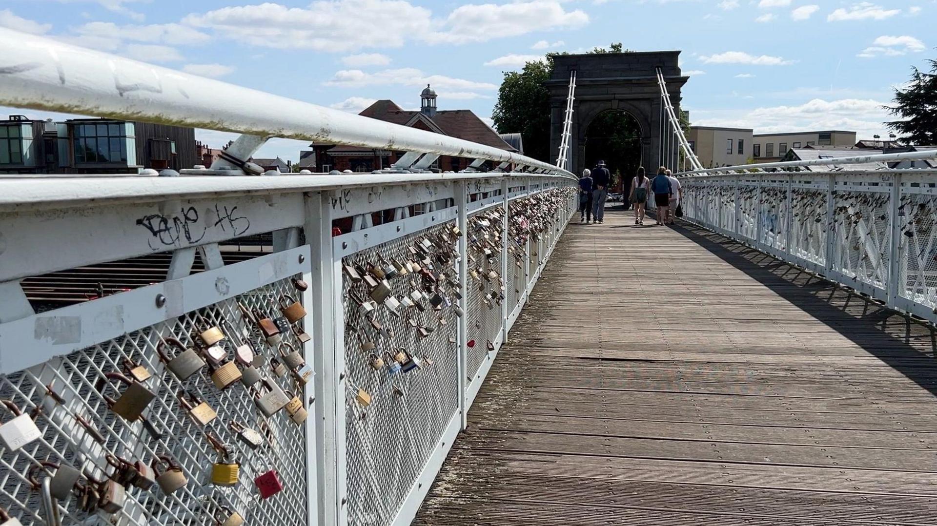 Wilford Suspension Bridge with people walking on the bridge in the distance. Love locks can be seen attacked to a the bridge on either side. 