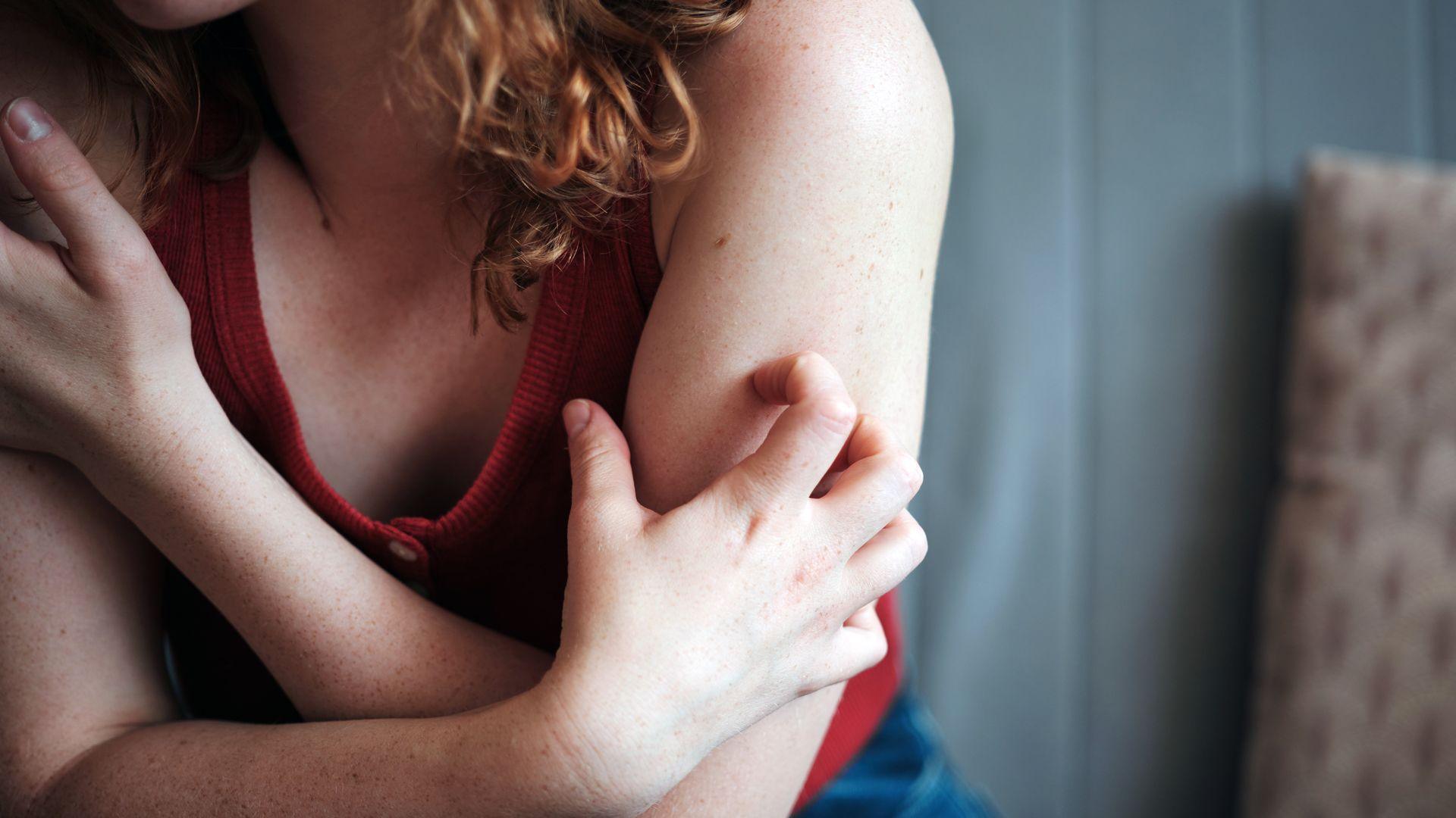 A young woman, wearing a dark red top but with bare arms, anxiously digs her fingers of one hand into the top of her arm. The woman, whose face is out of shot, has long red curly hair.