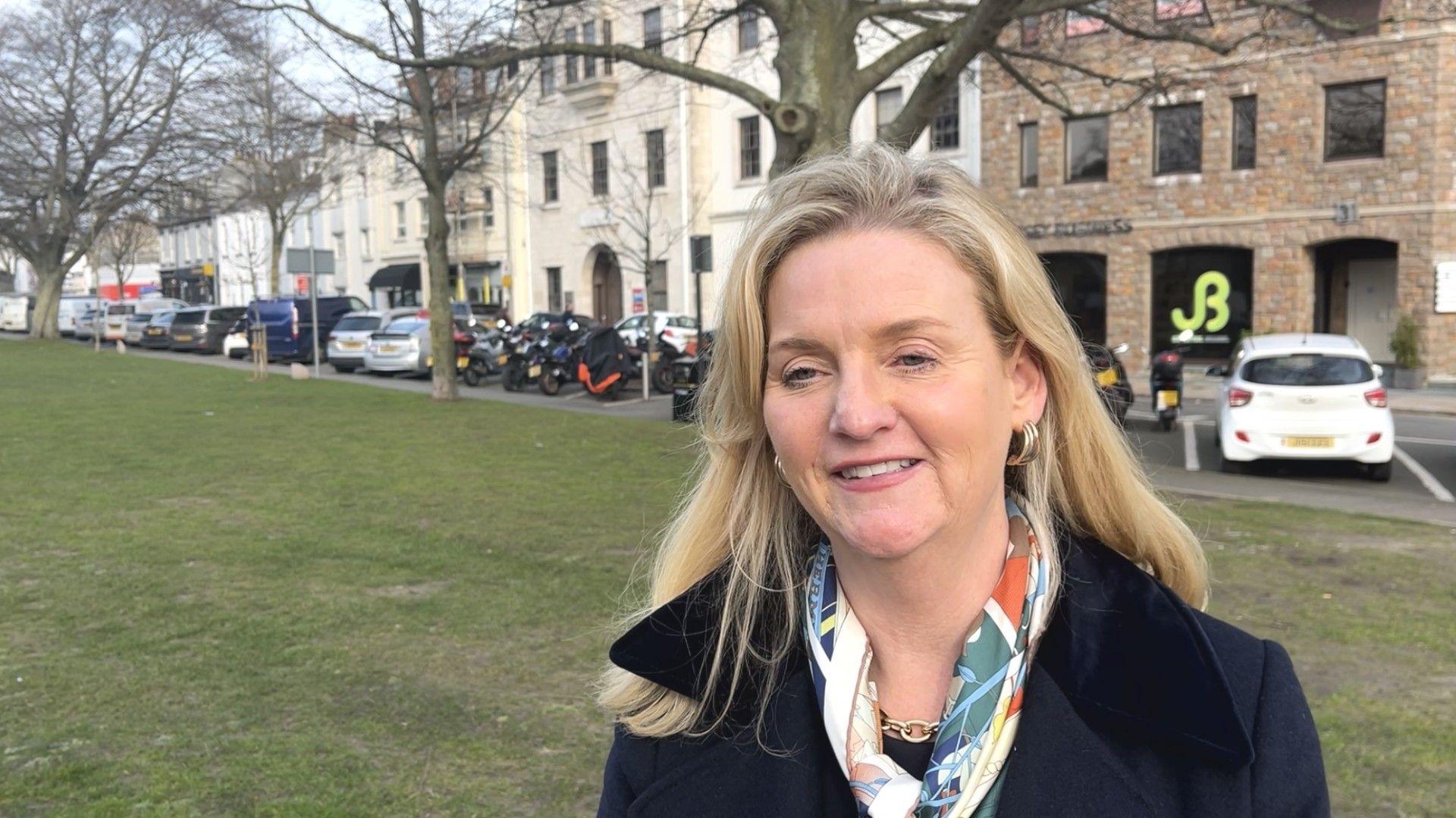 A blonde woman with long straight hair standing in a park wearing a navy blue coat and colourful scarf. Parked cars and buildings are visible behind her.