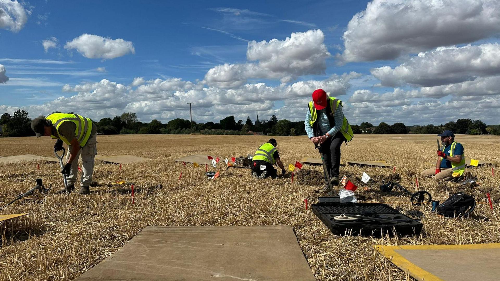 Four people are digging or examining a field, which is brown and stubbly. They are wearing caps and hi-vis yellow jackets. It is a sunny day and the sky is blue with billowing white clouds