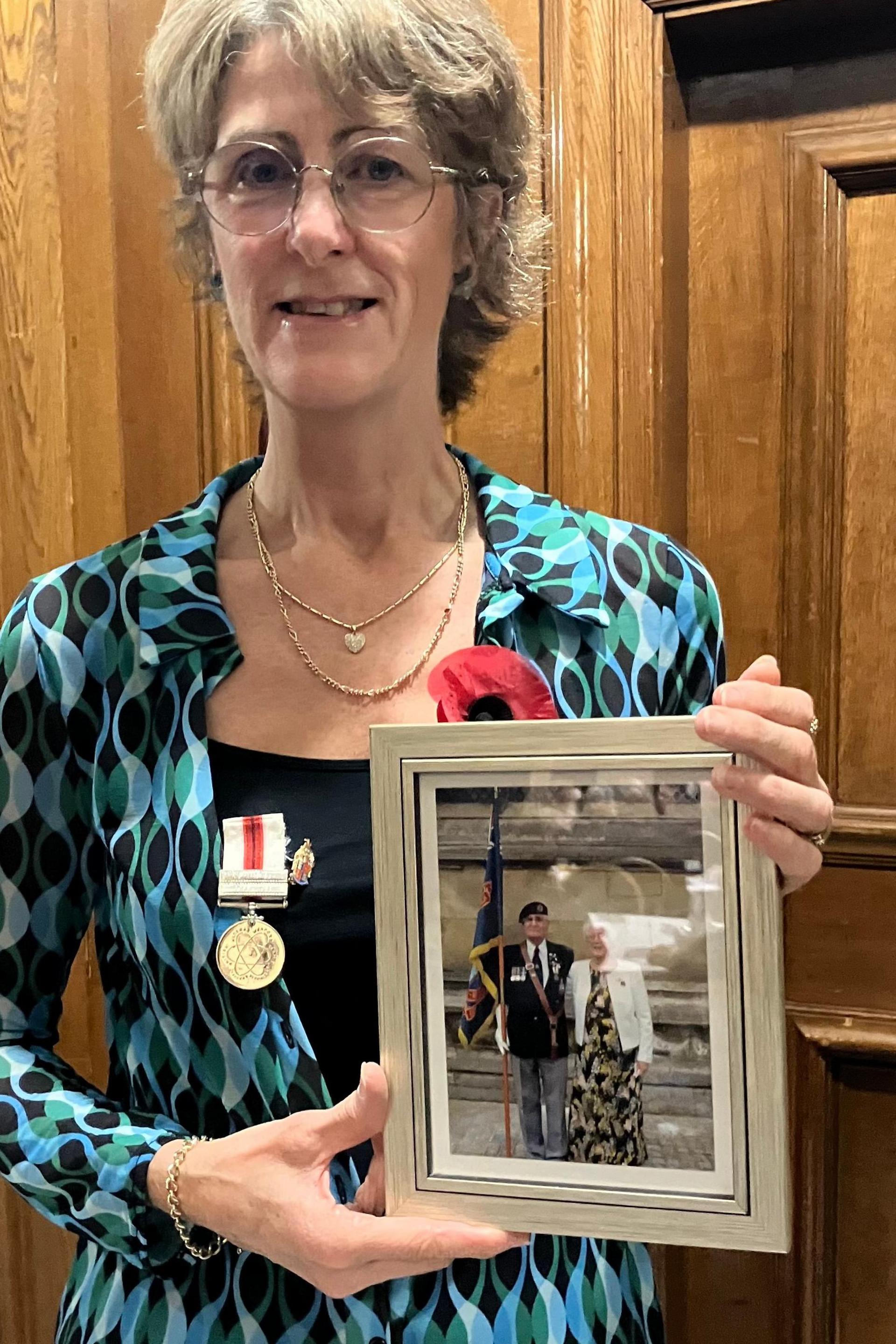 A woman with short blonde hair standing indoors holding a framed photograph showing two individuals in formal attire, one wearing a military-style uniform and the other in a light jacket and patterned skirt. She wears glasses, a blue top, a black vest and a medal and red poppy.