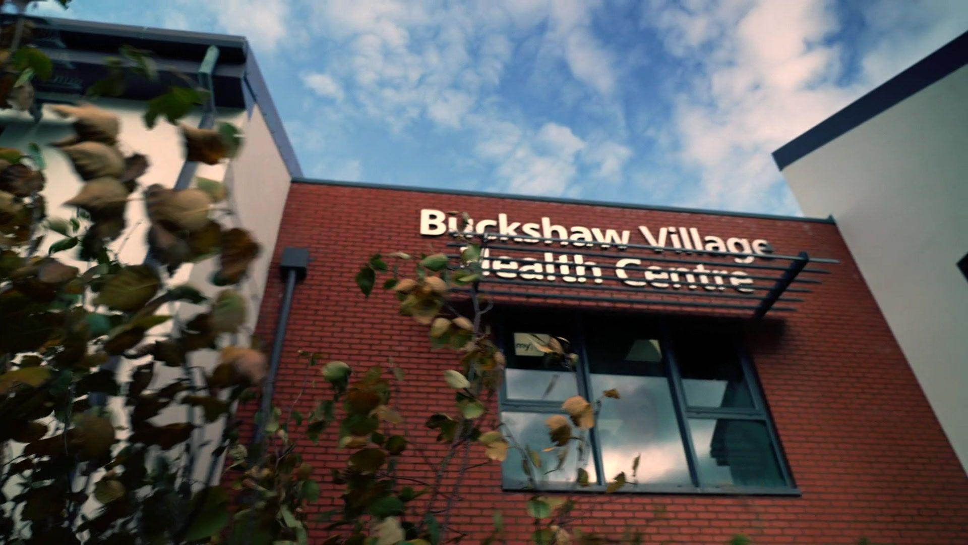 An external view of the Buckshaw Village Health Centre building with red brick and white rendered walls set behind green shrubs.
