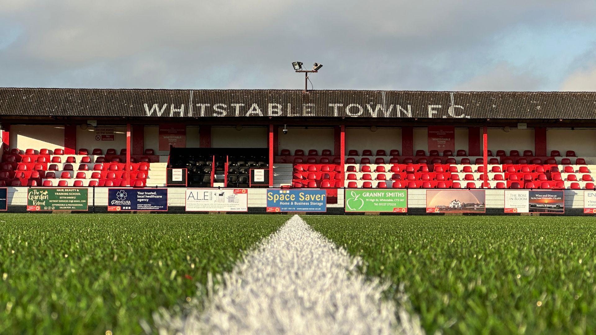 A photograph of a football stand with WHITSTABLE TOWN F.C. written on the roof. The photo is taken from grass level on the other side of the pitch