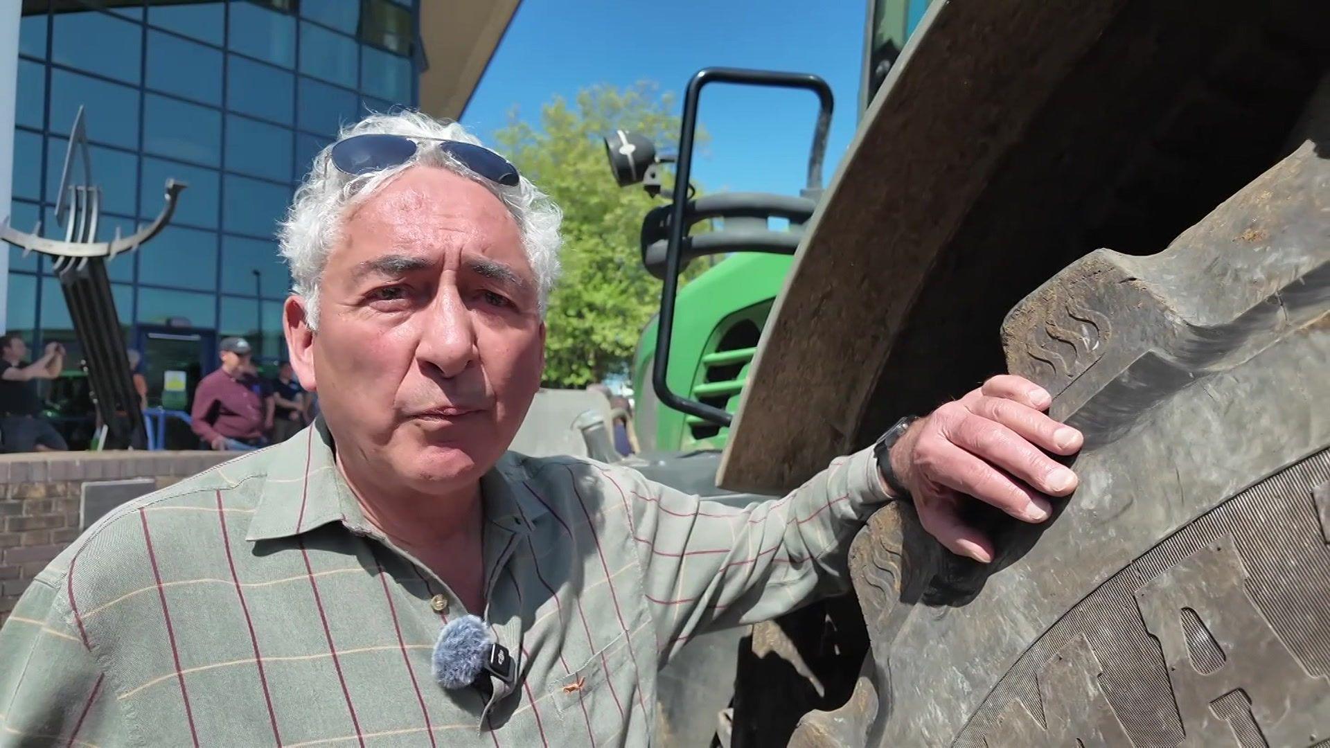 Mark stands next to a tractor giving a TV interview. He has curly, white hair with sunglasses over his head and wears a checked shirt.