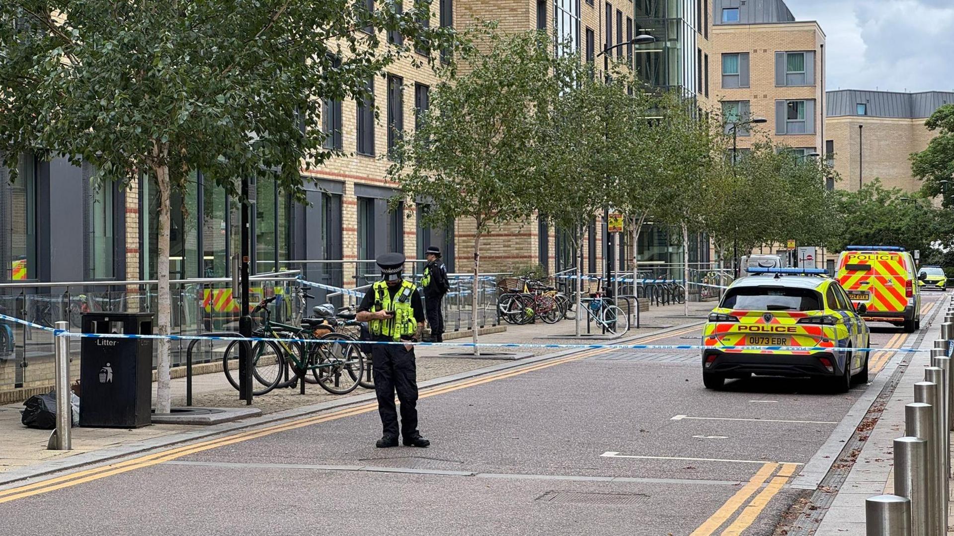 A street is cordoned off by blue-and-white tape. A police car and police are van parked on the road. Two officers can be seen within the cordon, which is on a tree-lined street with modern buildings.
