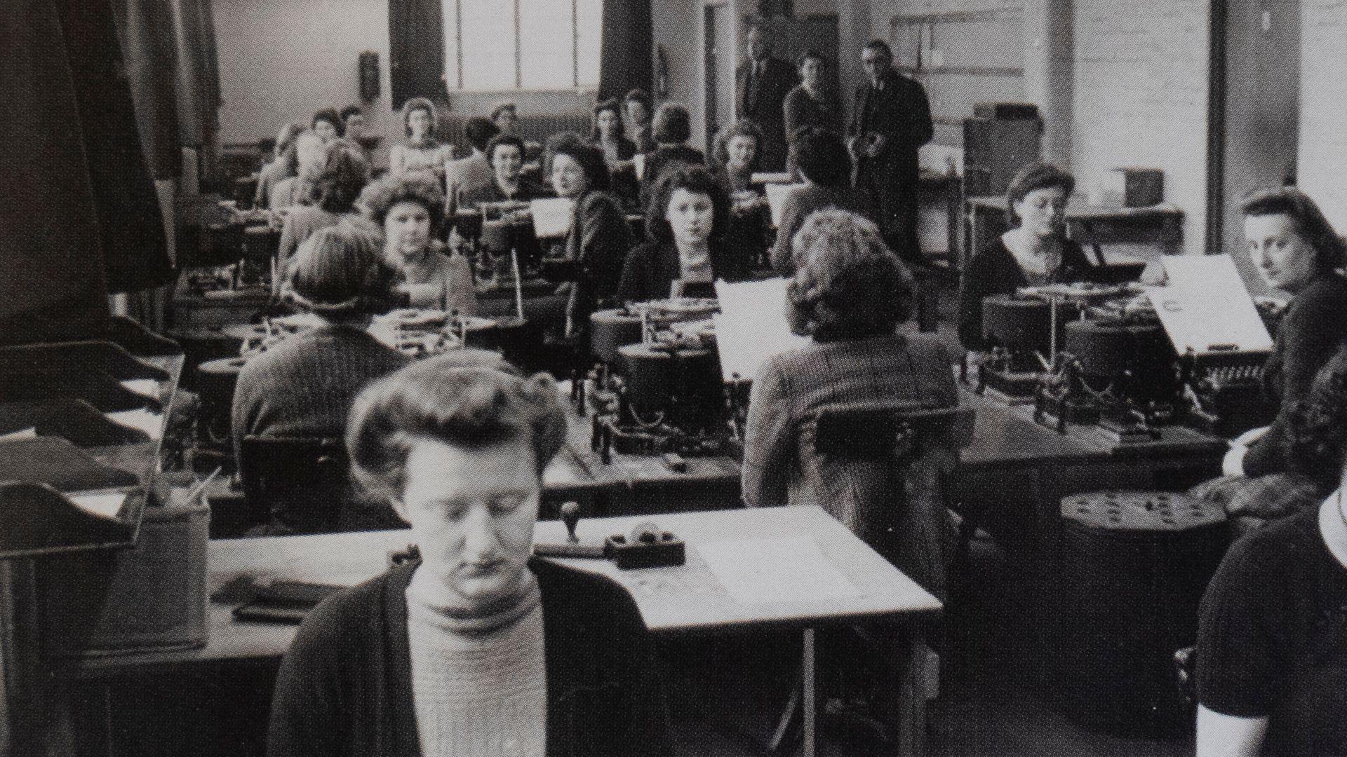 A black and white photo of a group of women sat at desks working at Bletchley Park during the war. You can see machines in front of them which they are using to crack codes.