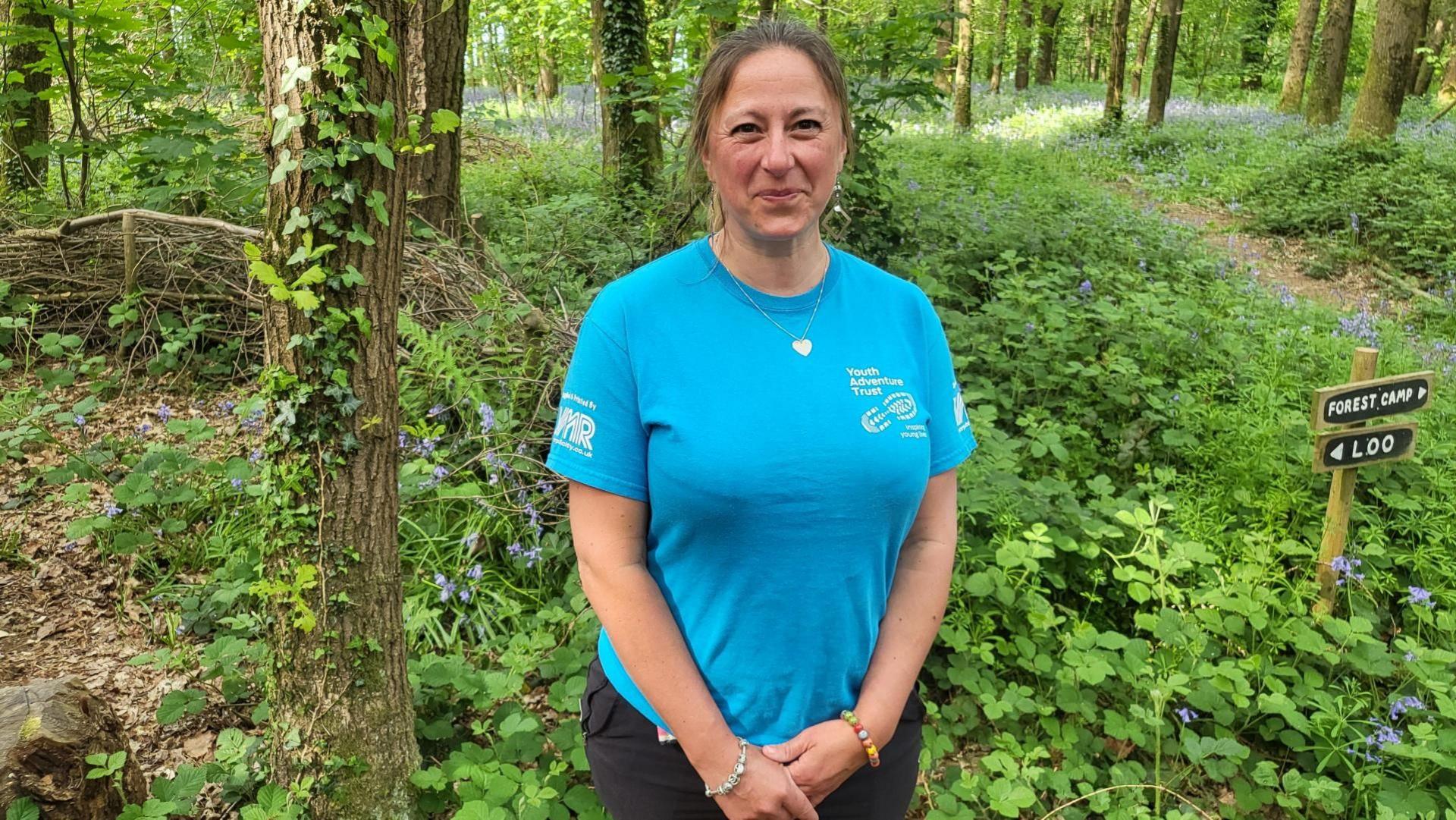 Louise Balaam is wearing a blue Youth Adventure Trust t shirt and some colourful beaded bracelets. She is smiling at the camera and there is a forest camp and loo sign behind her, amid trees and woodland.