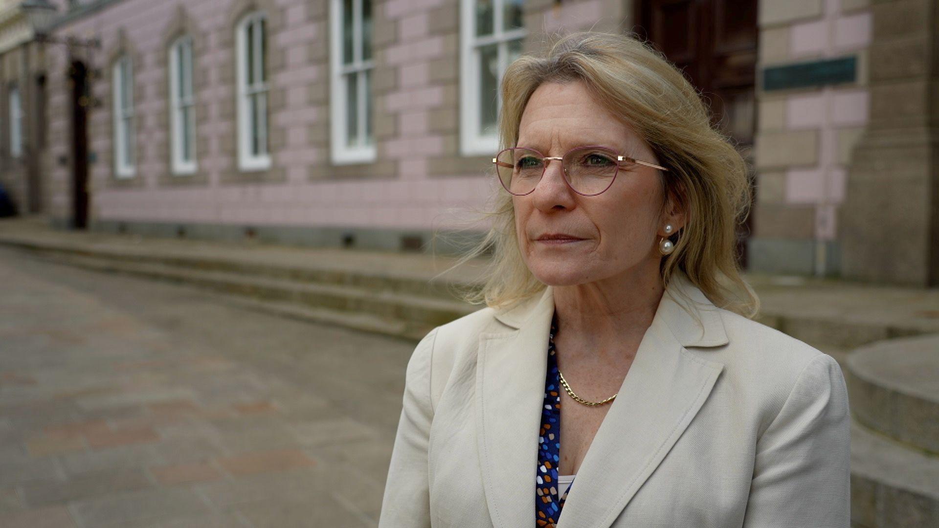 Treasury Minister Elaine Millar in Jersey's Royal Square. She has shoulder-length blonde hair, circular metal glasses and is wearing a cream formal jacket