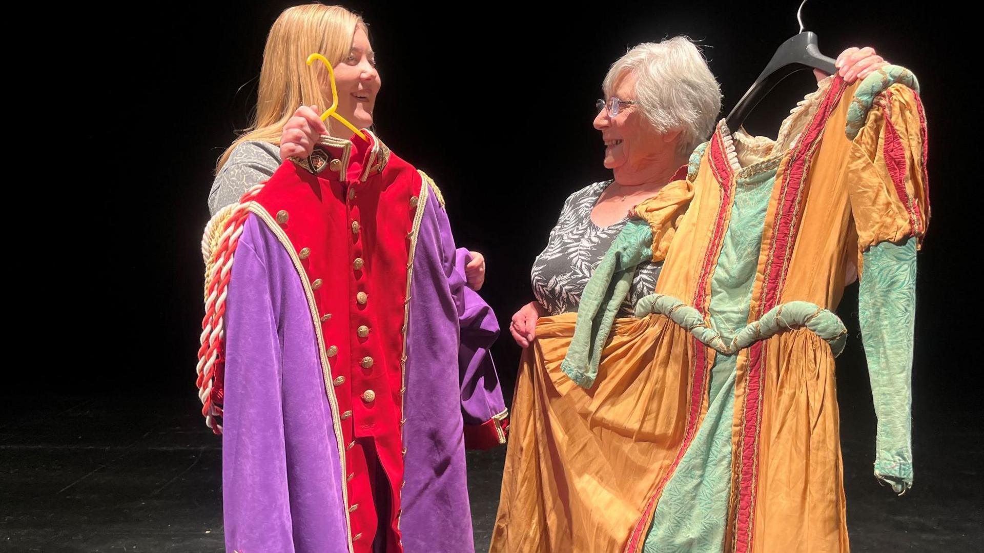 Two smiling women stood on a stage looking at each other. Both women are holding elaborate theatrical costumes. The woman on the left has long blonde hair and is holding a long, regal-looking coat in purple and red with gold trim and buttons. The other woman has short, light grey hair and is wearing glasses. She is holding a golden gown with green sleeves and red detail. The background is dark.