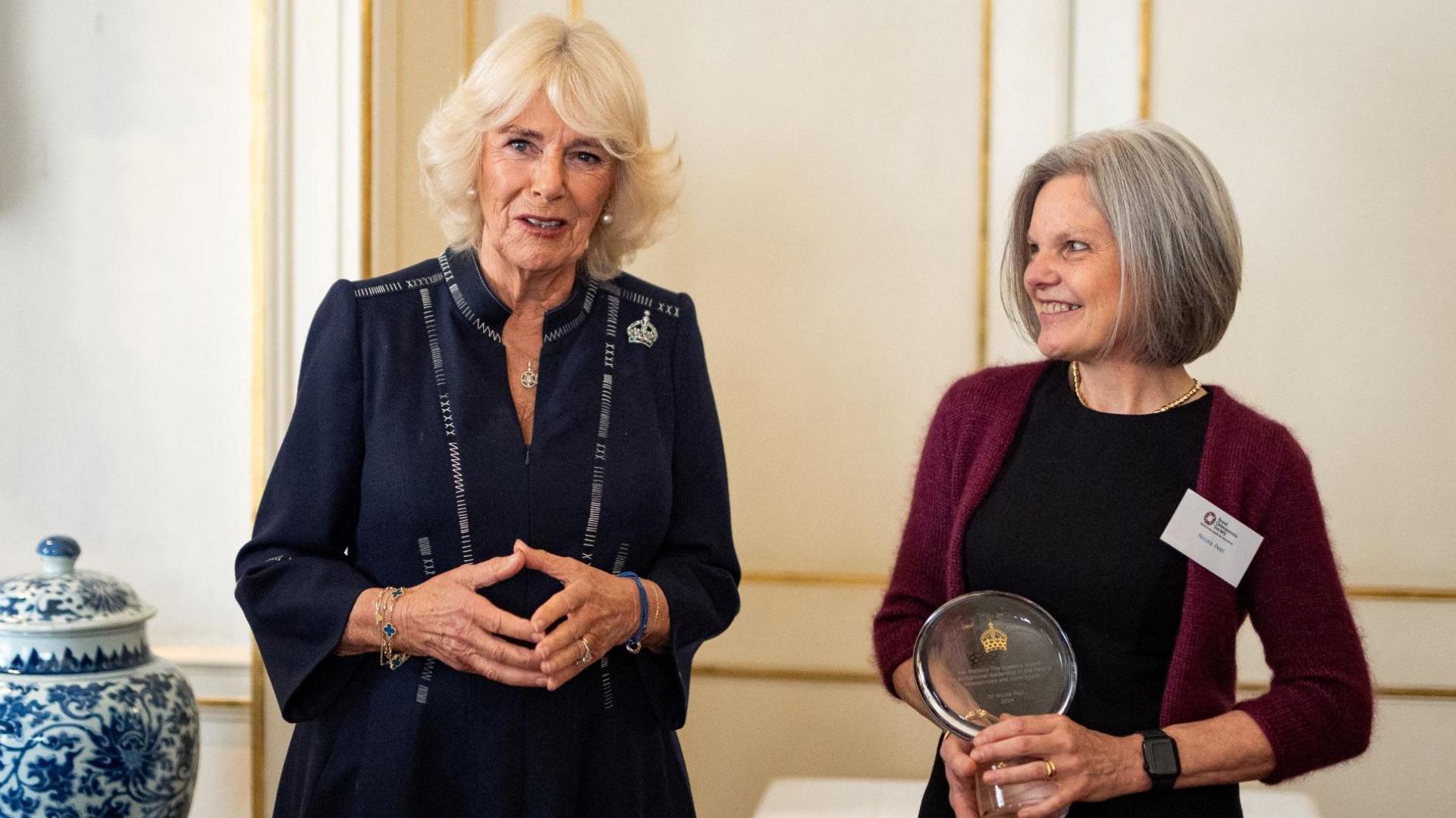 Queen Camilla wears a navy dress with a zipper on the front. She has her fingertips pressed together and smiles at the camera. Dr Peel is wearing a black dress and burgundy cardigan. She holds the glass award and looks at the Queen with a smile.